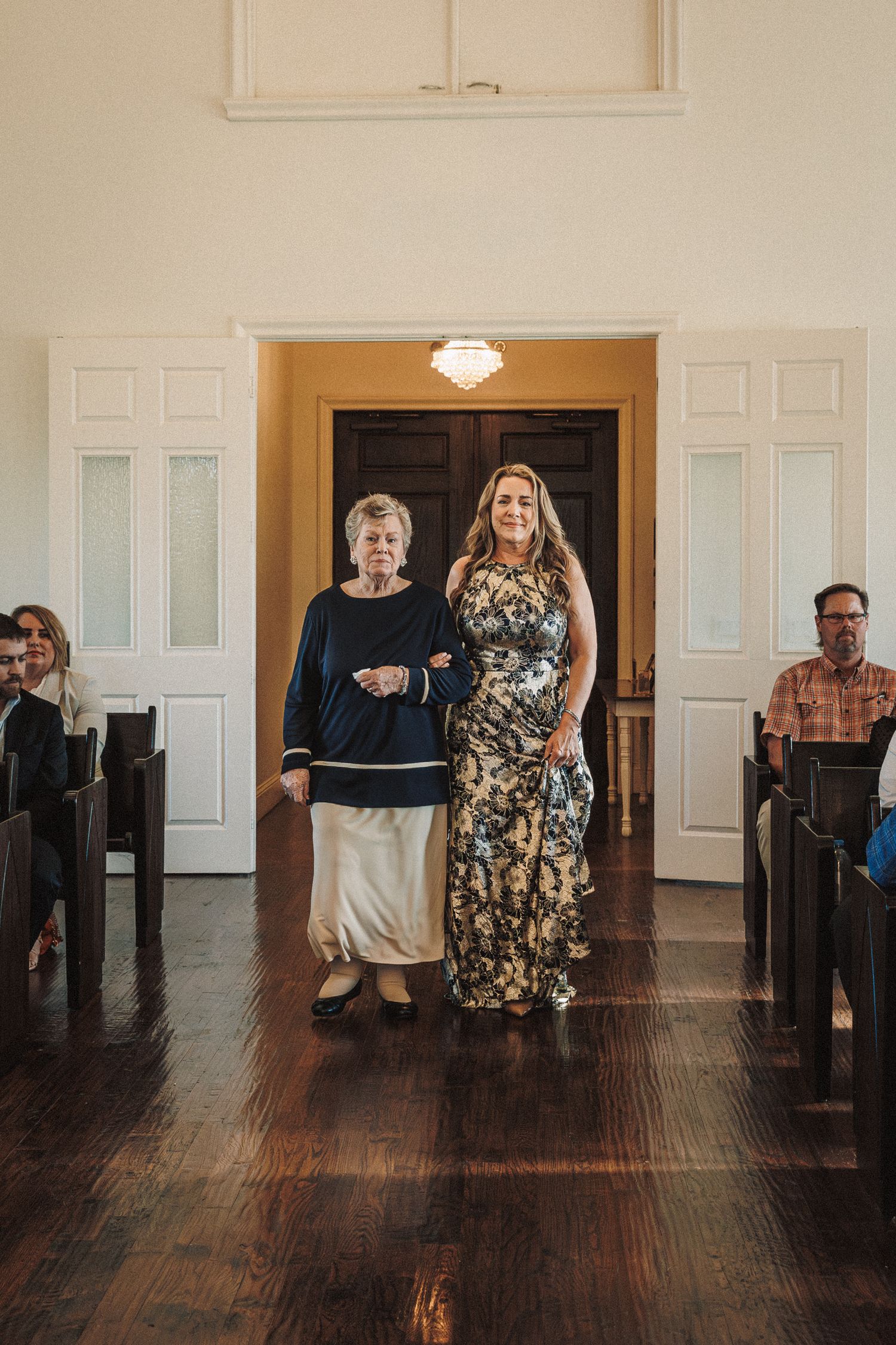 Two women walk arm-in-arm down a church aisle during a wedding ceremony.