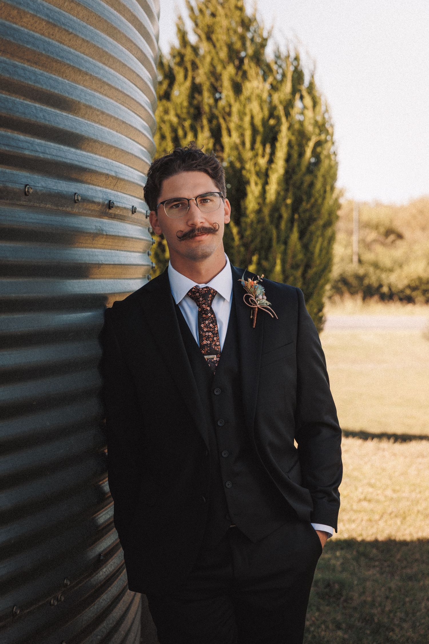Groom in a black suit with floral tie and boutonniere leaning against a metal grain silo.