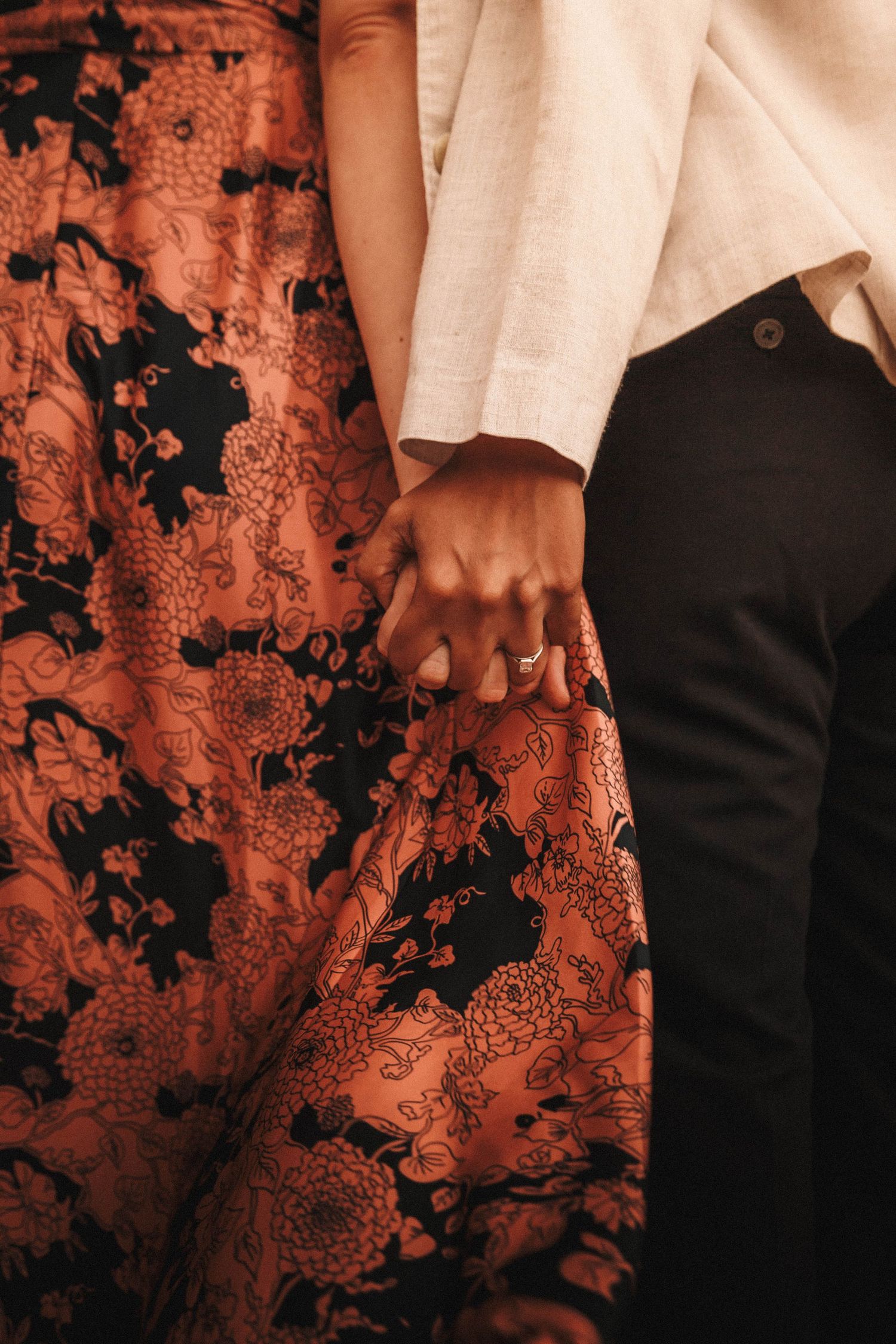 Close-up of engaged couple holding hands, showing engagement ring, she wears a floral orange and black maxi skirt.
