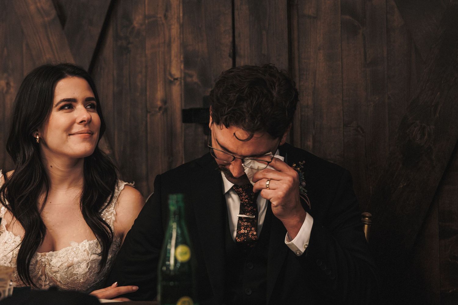 Bride smiles warmly as emotional groom wipes tears during wedding reception at rustic barn.