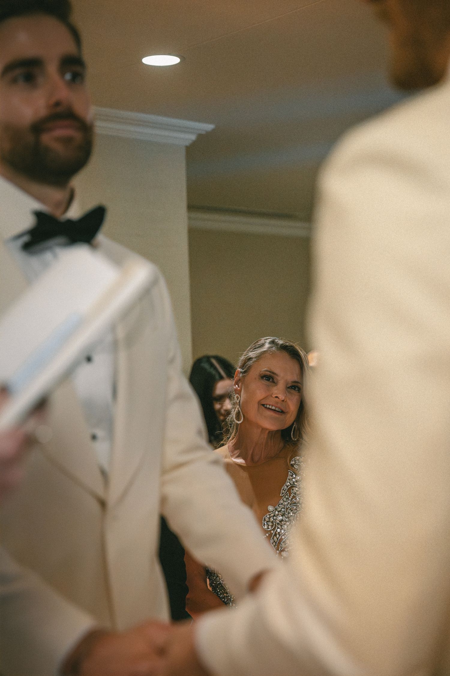 A smiling woman in a beaded gown watches two grooms exchange vows at their wedding ceremony.