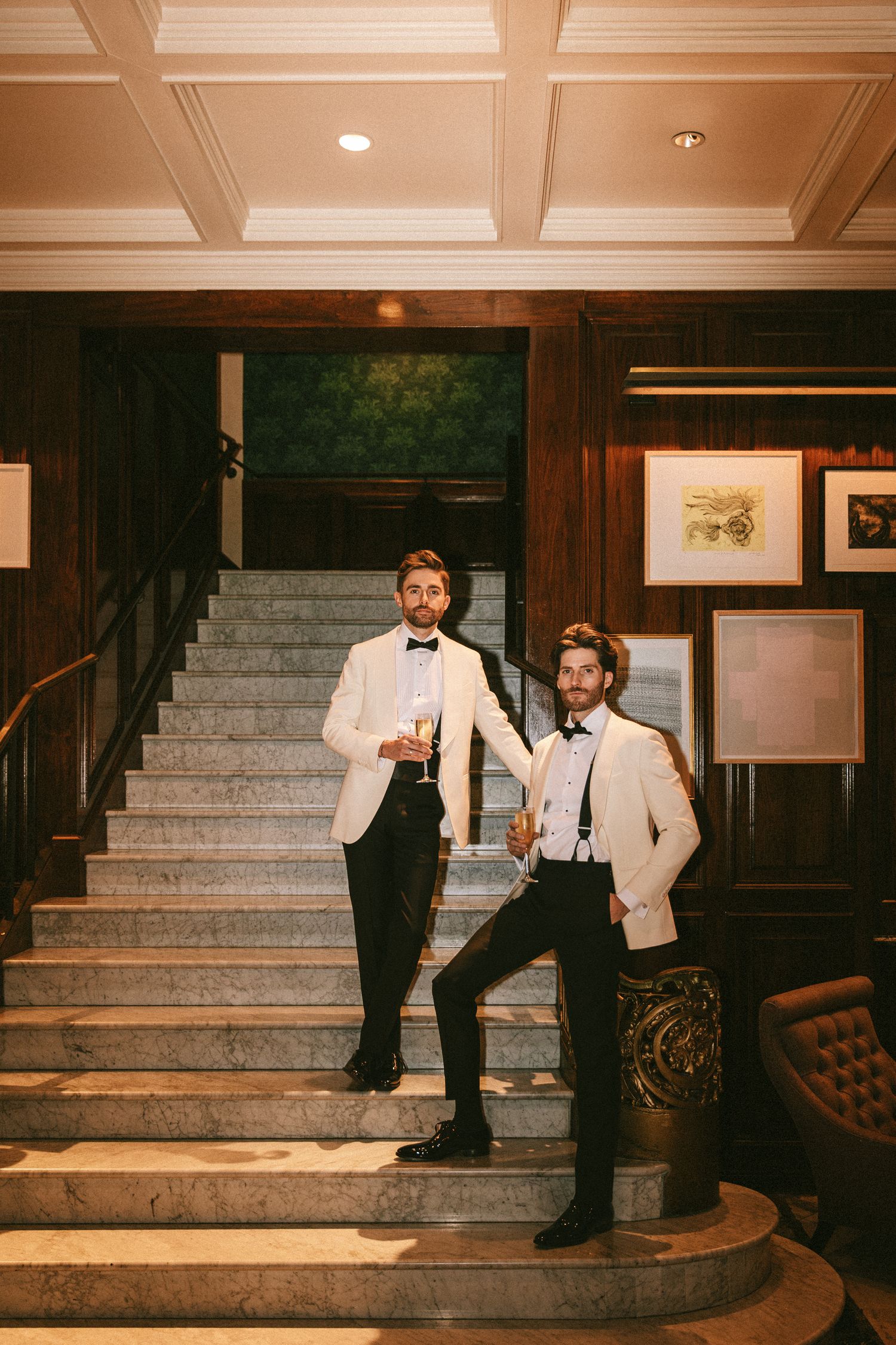 Two grooms in matching ivory tuxedos pose confidently on grand marble staircase holding champagne glasses.