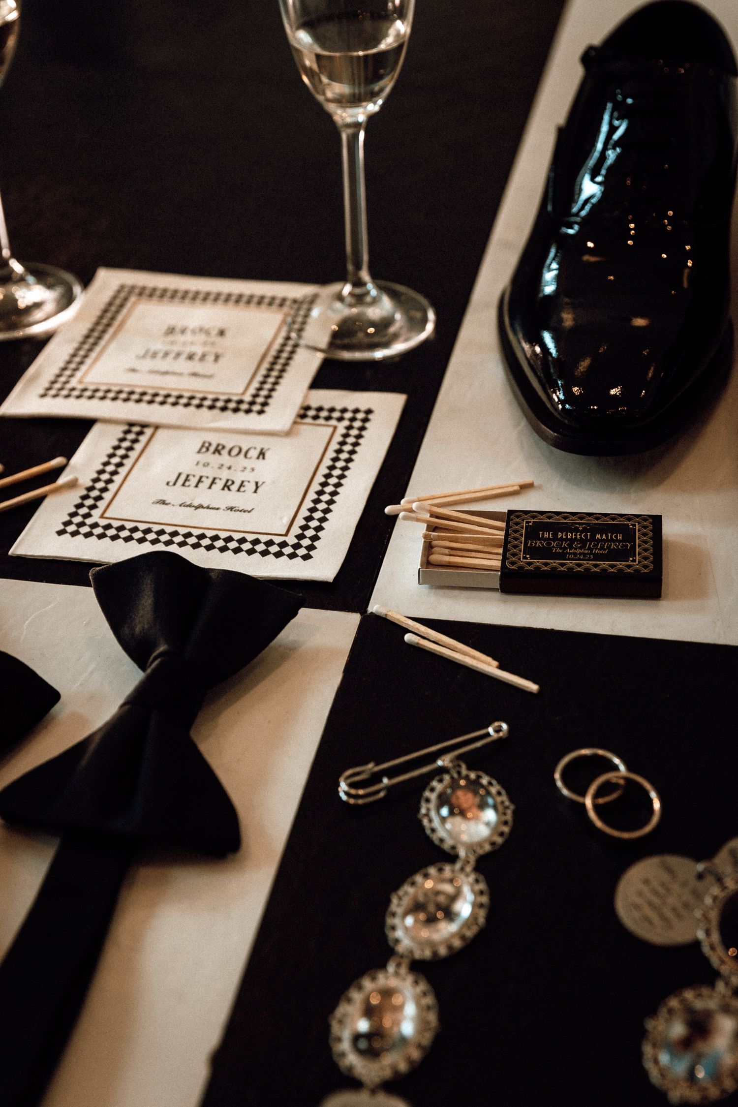 Black and white wedding stationery with houndstooth border, bow tie, rings, brooches, matchbox, and patent leather shoe on table.