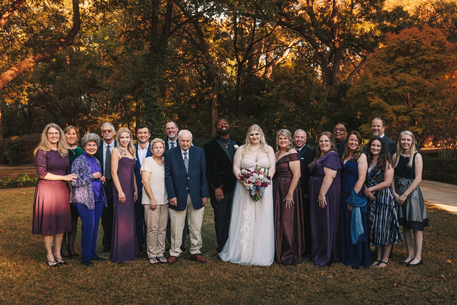 Bride and groom posing with large wedding party in purple dresses amid autumn trees.