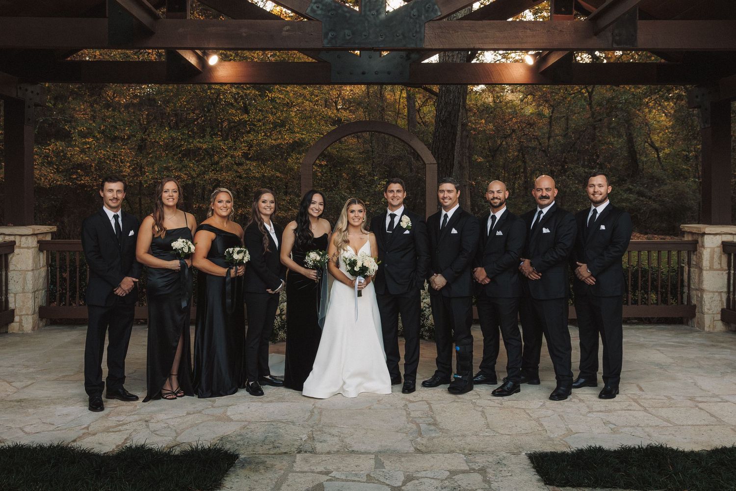 Wedding party in black attire with bride and groom posing under rustic pavilion.
