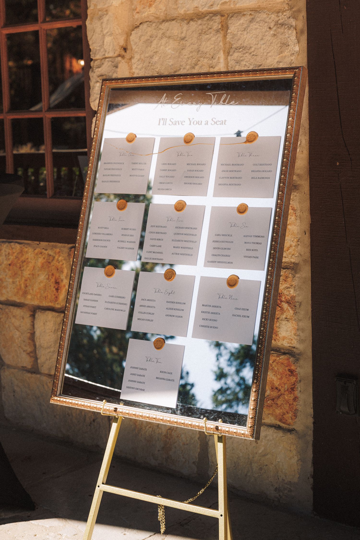 Wedding seating chart displayed on ornate gold mirror frame on easel with wax seal accents reading I'll Save You a Seat.