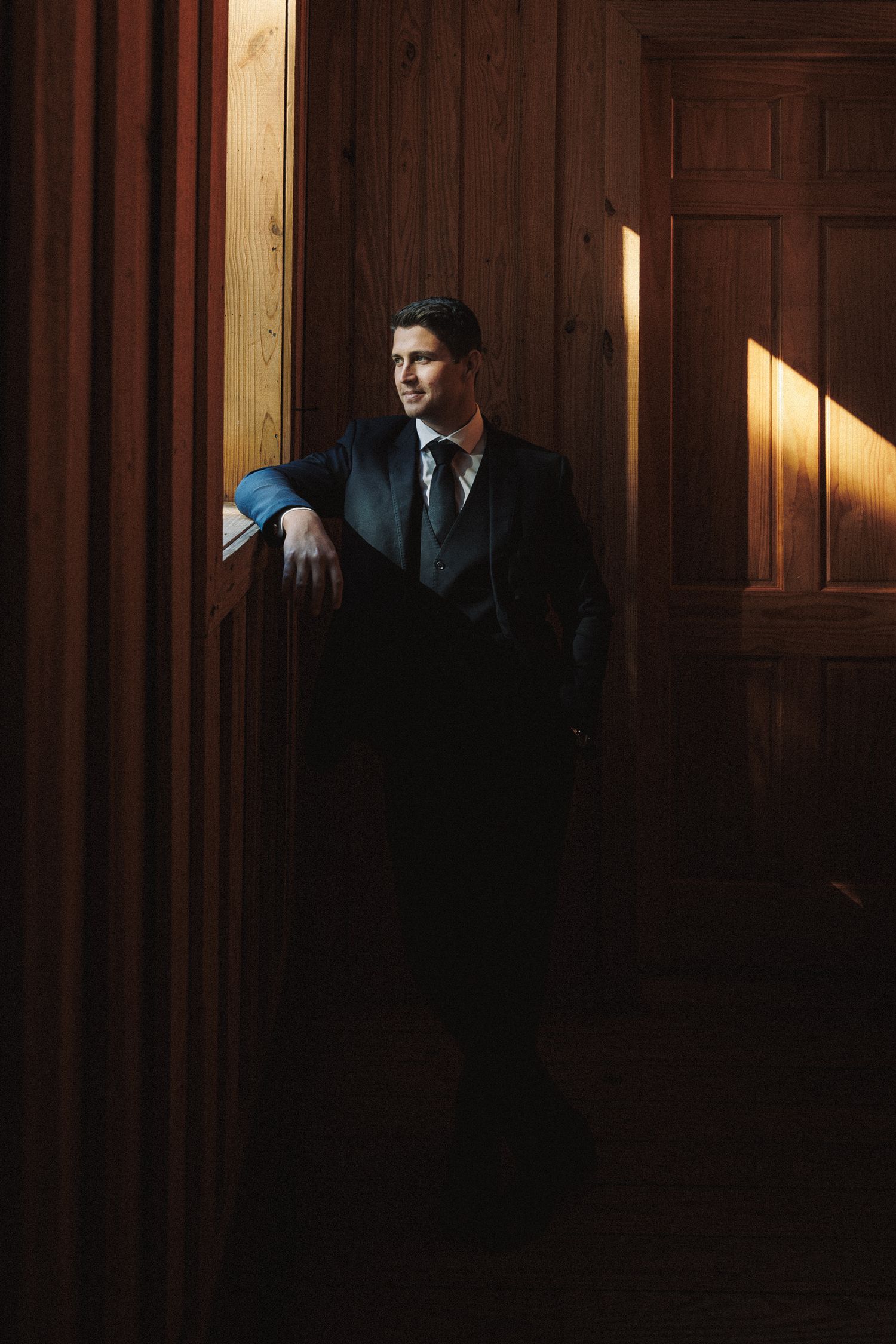 Groom in dark suit and tie leaning against wooden wall in dramatic moody lighting.