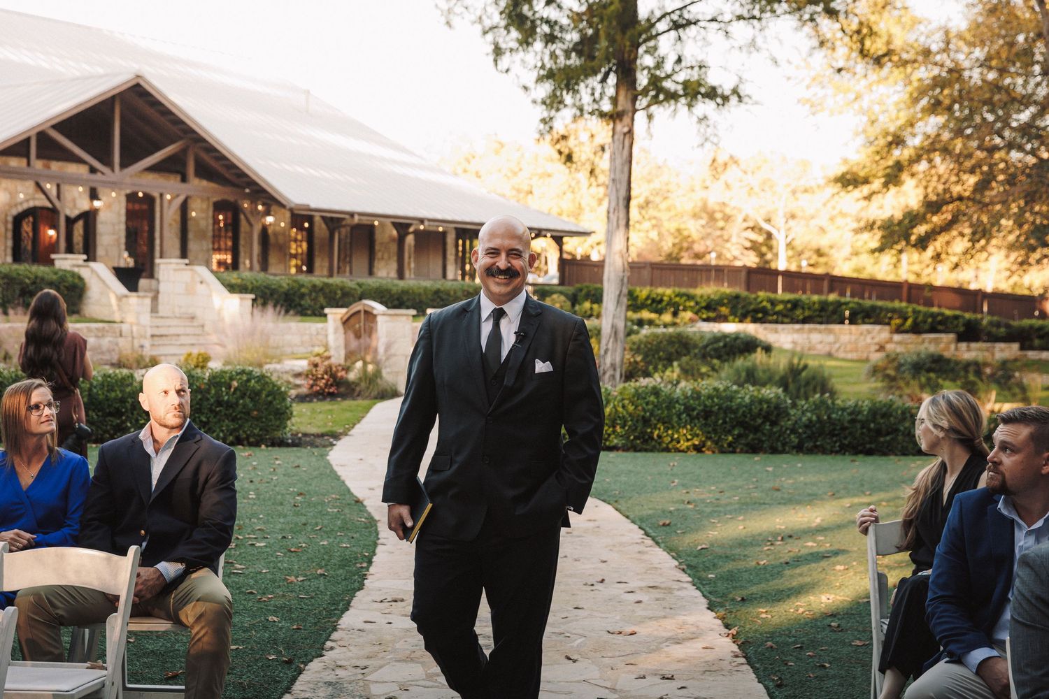 Smiling man in black suit walks down outdoor wedding ceremony aisle at sunset.