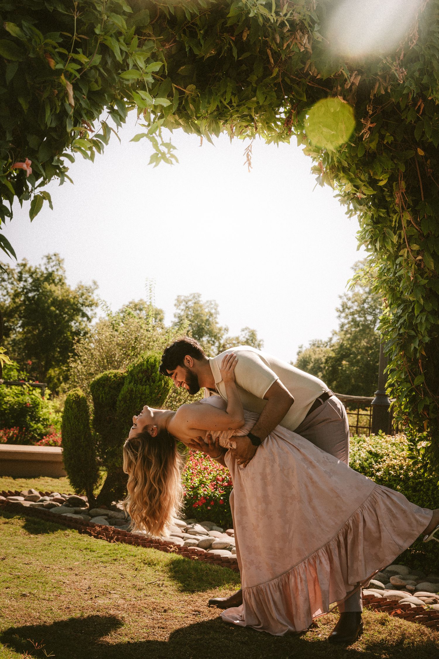 A romantic couple embraces in a sun-drenched garden setting during golden hour with lush greenery surrounding them.