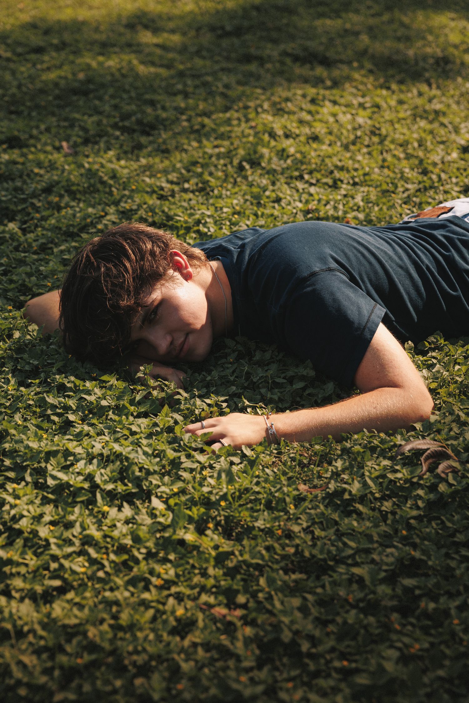 A person lies peacefully in a bed of lush green ivy or ground cover in a garden setting during daytime.