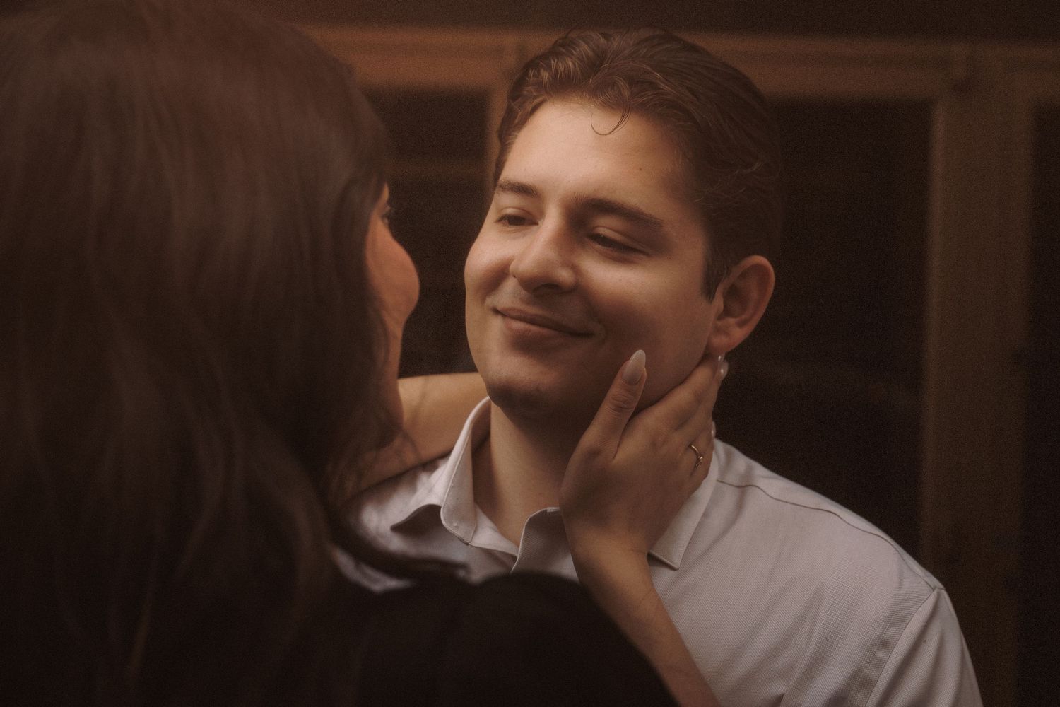 A woman tenderly touches a smiling man's face in a warm, intimate moment in a dimly lit room.