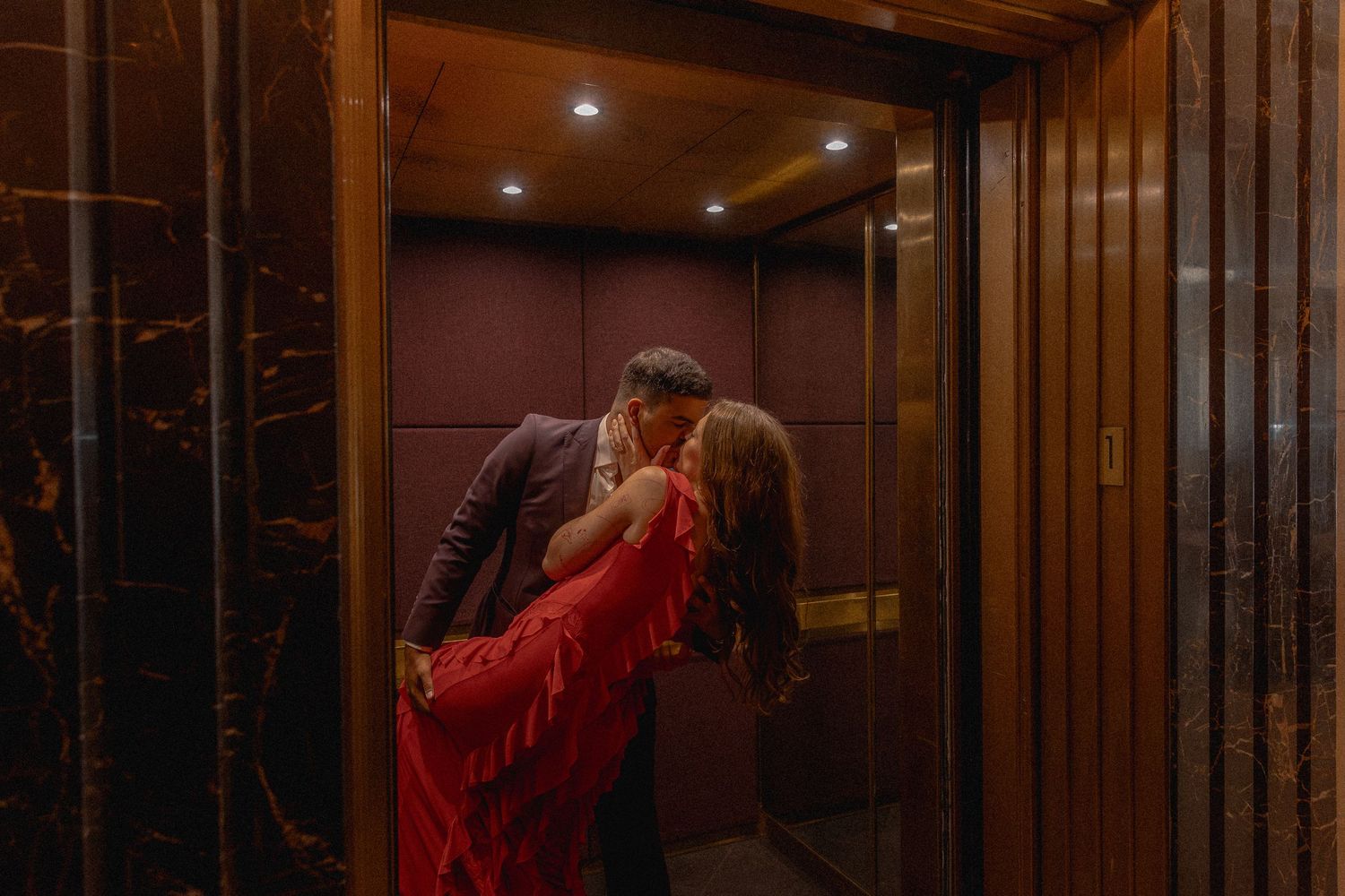 Couple sharing a romantic kiss in an elegant elevator with marble walls and warm wood paneling, woman in red gown.