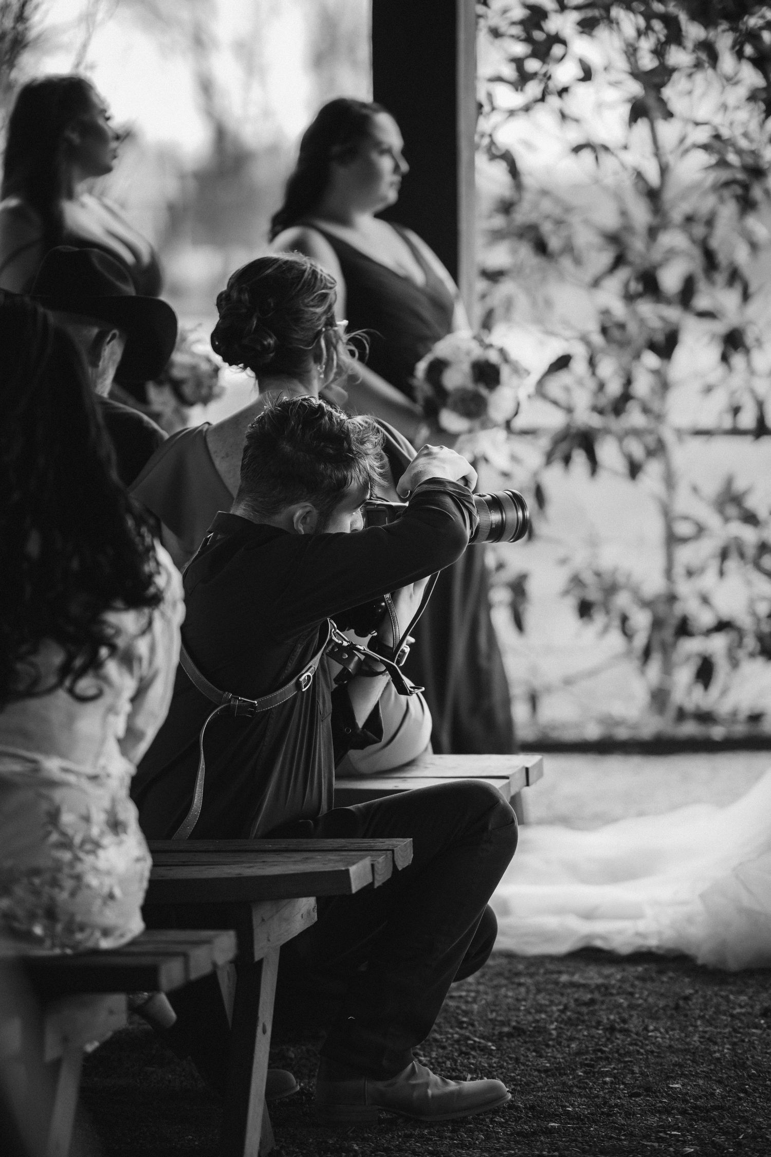 Black and white photo showing guests seated on wooden benches at an outdoor event with silhouetted foliage in background.
