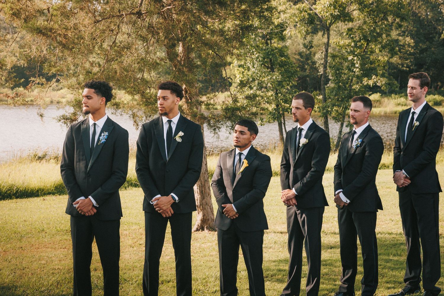 A wedding party with groomsmen in dark suits lined up at an outdoor venue.