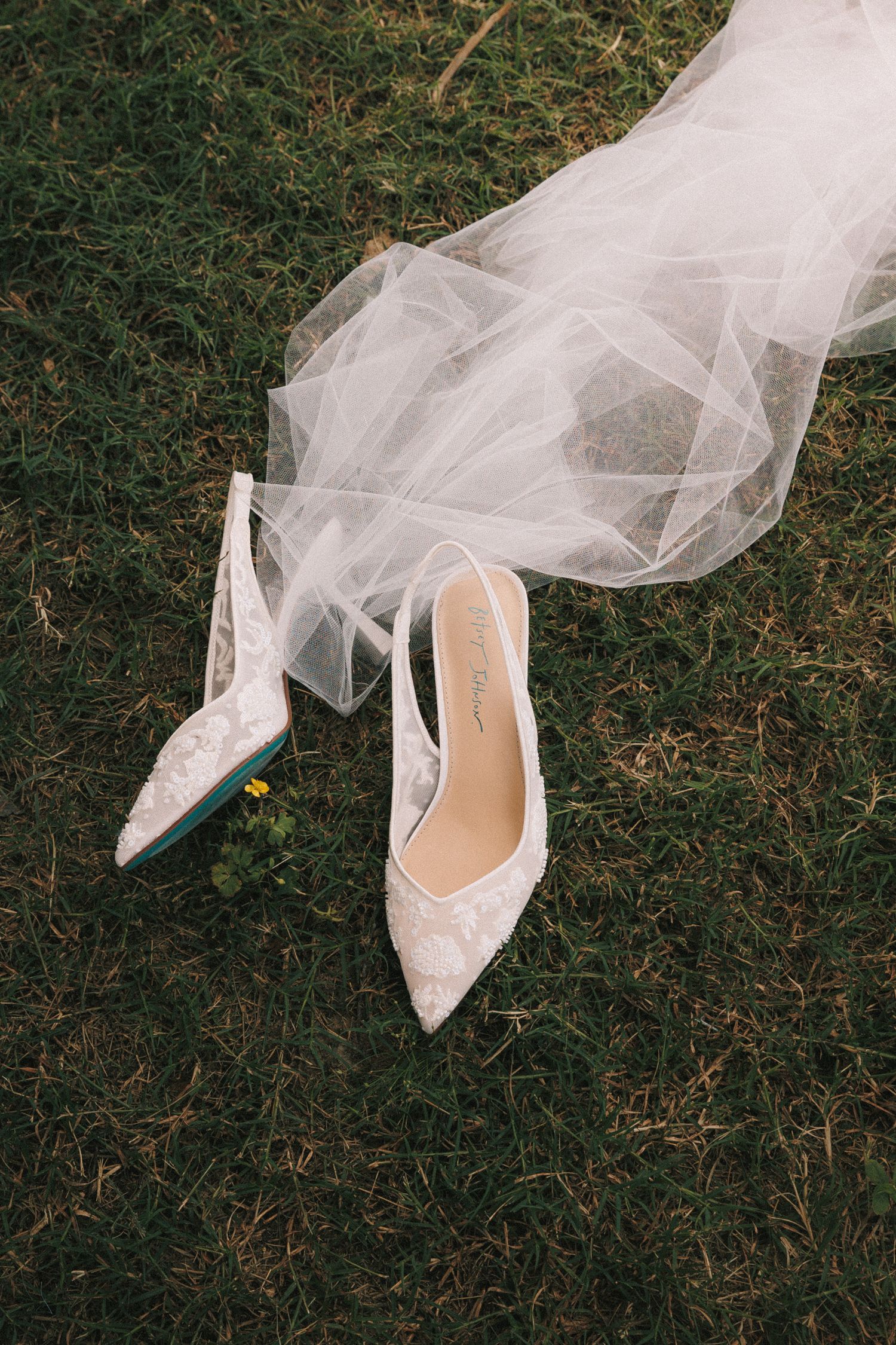 Delicate white lace wedding shoes with veil details on grass lawn.
