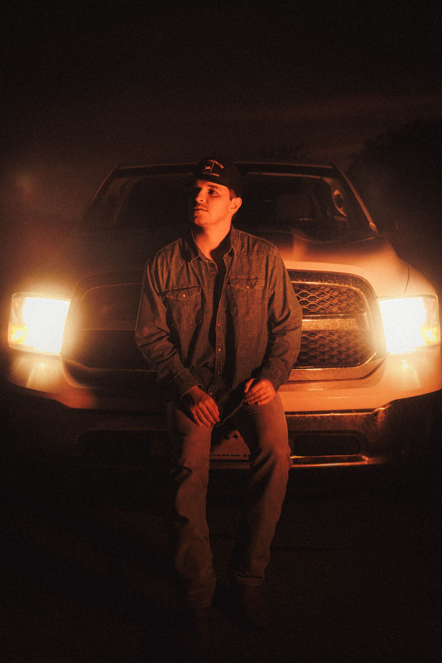 Silhouette figure in dark coat poses against car with illuminated headlights creating moody atmosphere at night.