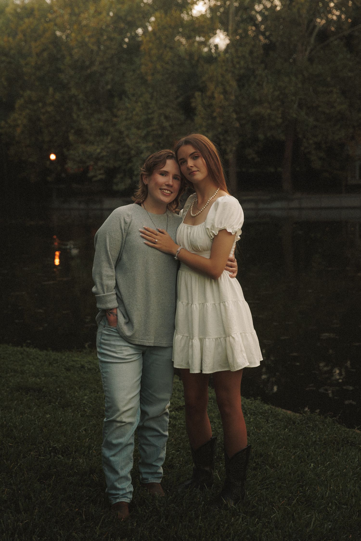 Couple in casual attire embracing outdoors during golden hour near a wooded area.
