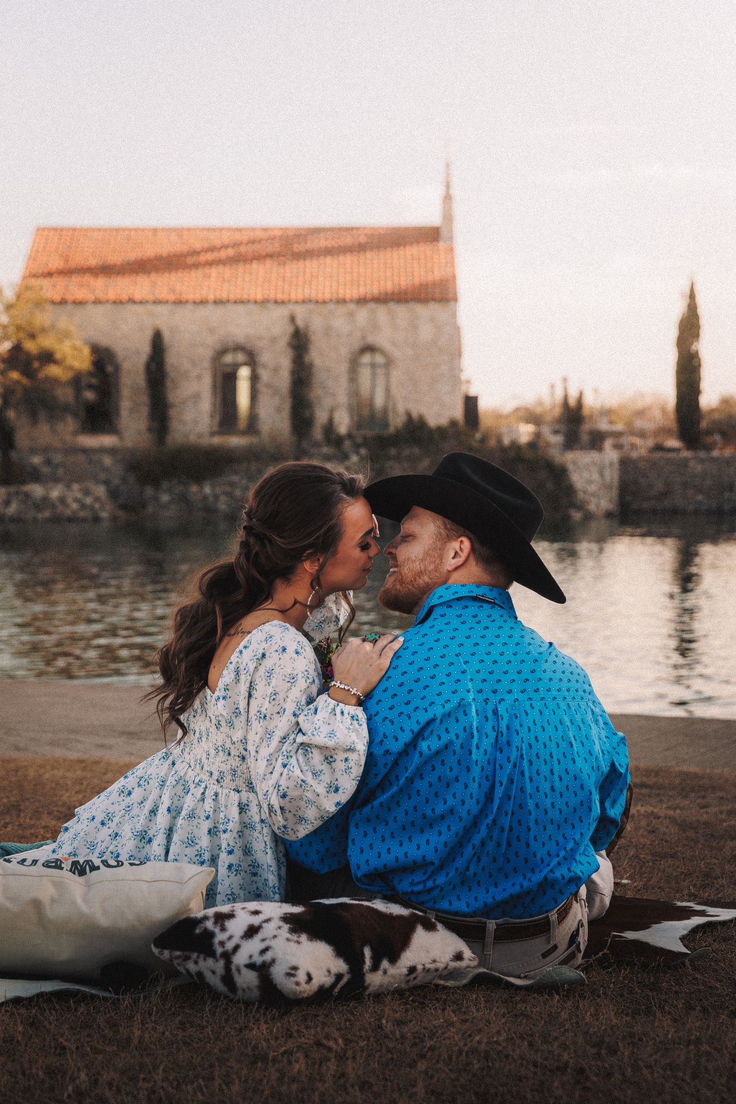 A romantic couple sits by a waterfront at sunset sharing an intimate moment against a historic European cityscape backdrop.