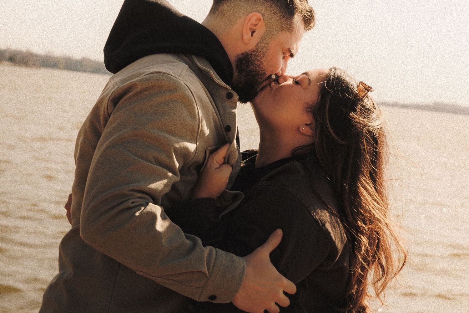 Close-up shot of a couple kissing tenderly by the water's edge in autumn clothing.