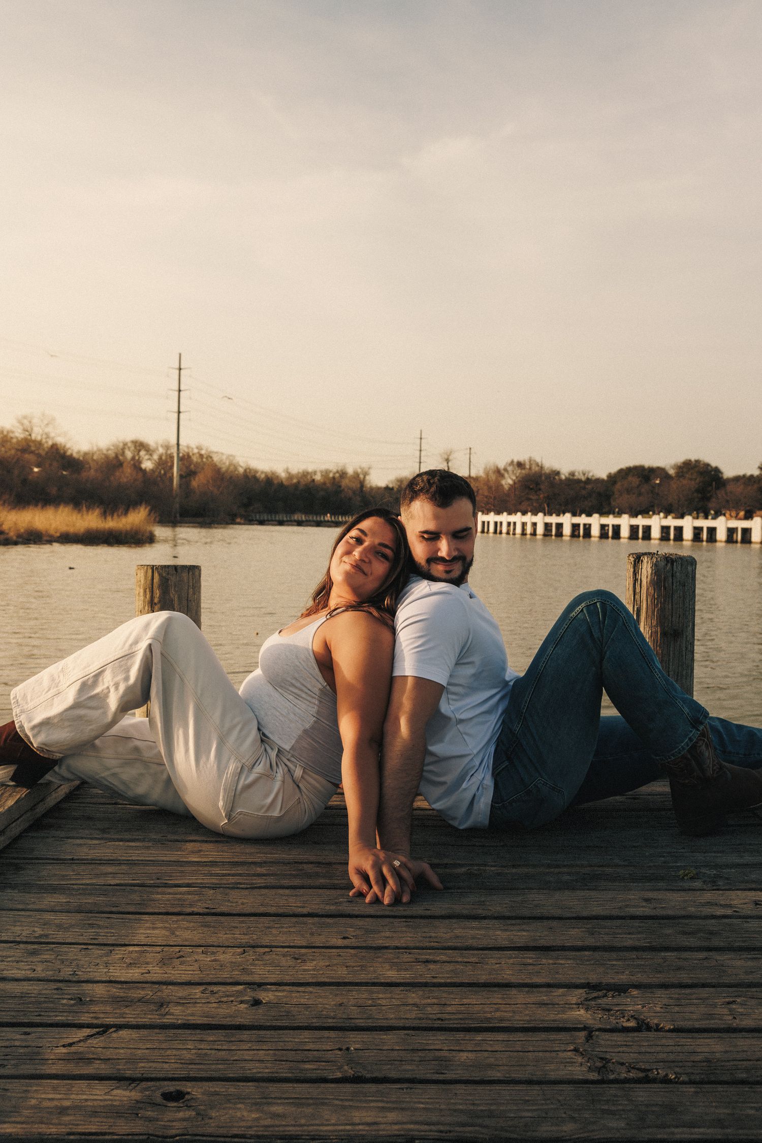 Couple relaxing back-to-back on wooden pier at sunset near water.