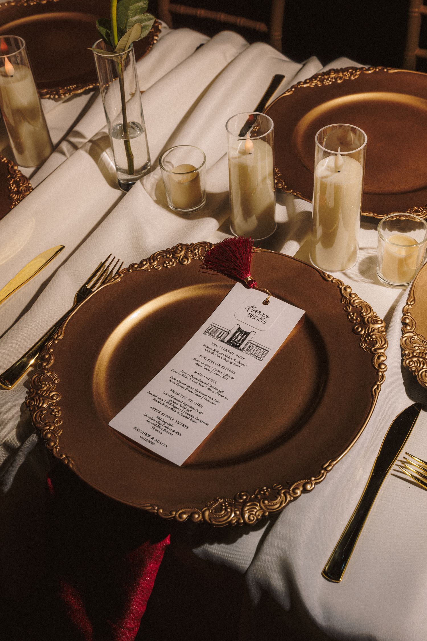 Elegant wedding table setting with gold charger plates, white menu card, candles, and gold cutlery on white linen.