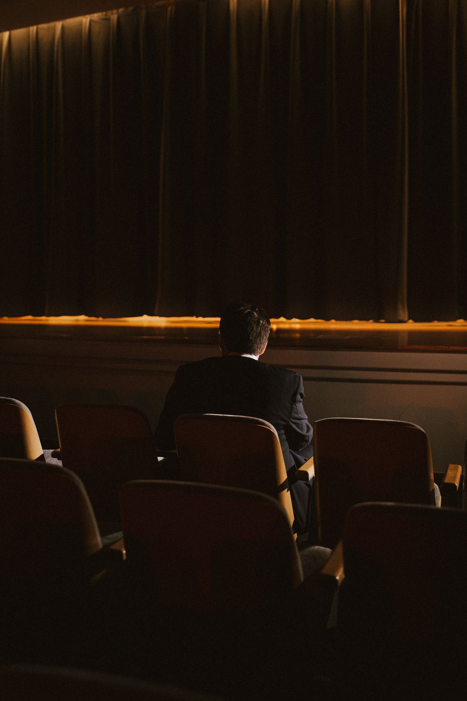 A man in a dark suit sits alone in a theater facing a large curtained stage in dim lighting.