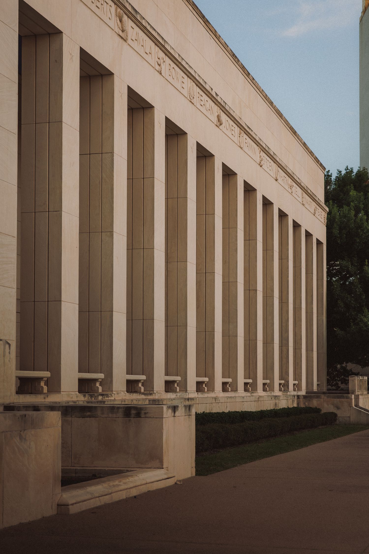 Classical neoclassical building with tall stone columns and manicured grounds under a blue sky.