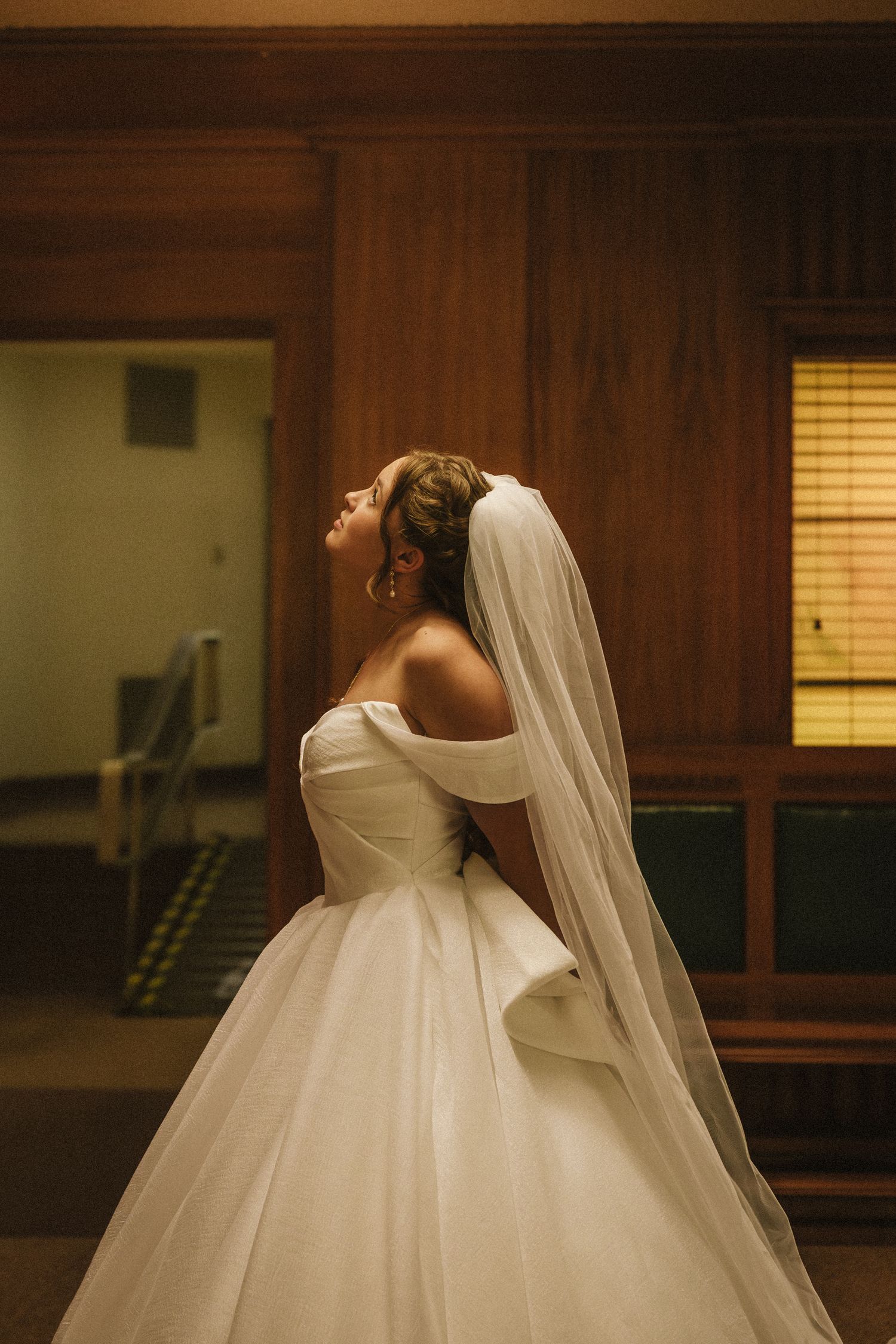 Elegant bride in off-shoulder ballgown with cathedral veil in warmly lit venue.