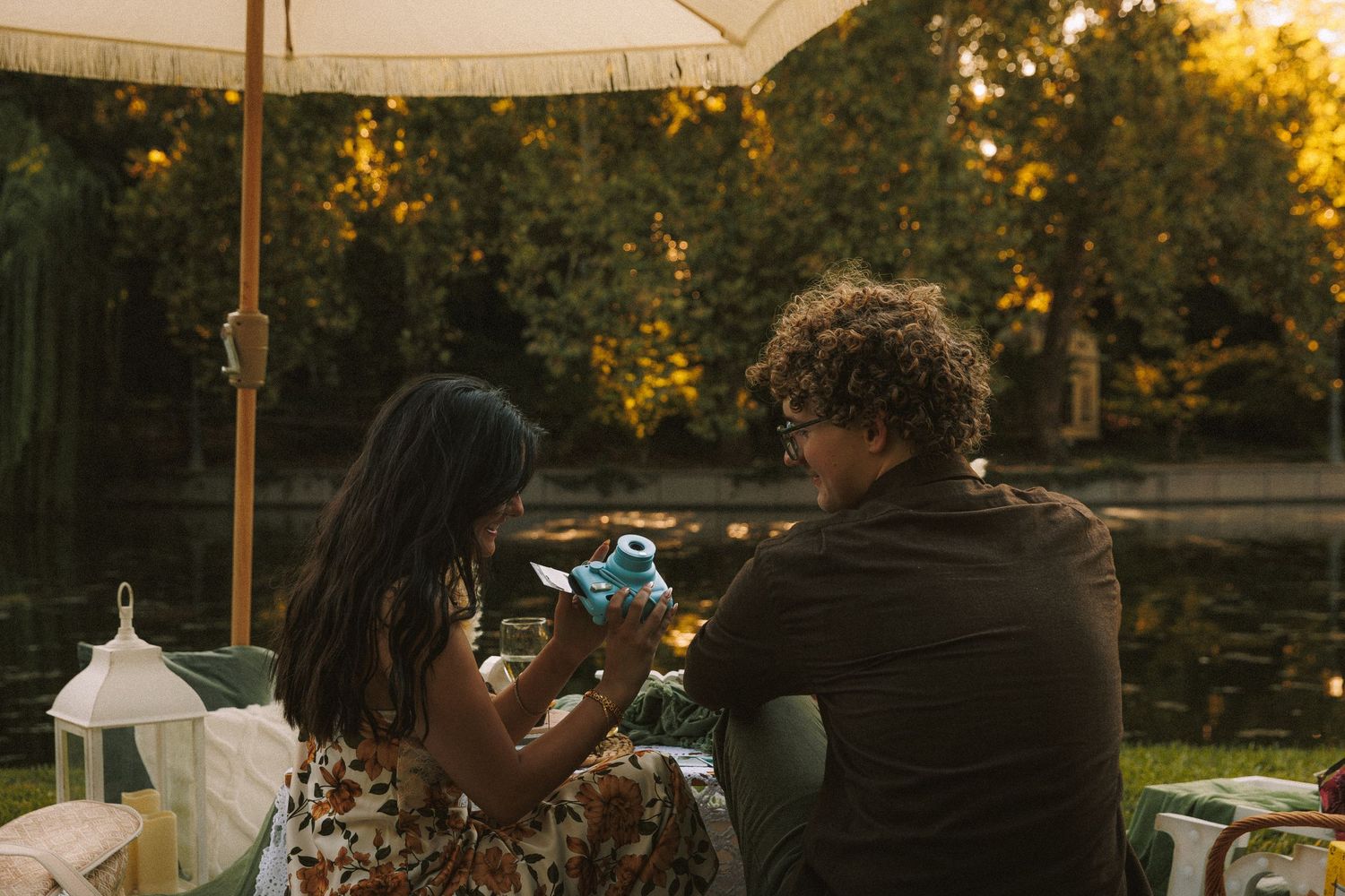 A couple shares drinks under an umbrella by a pond during golden hour.