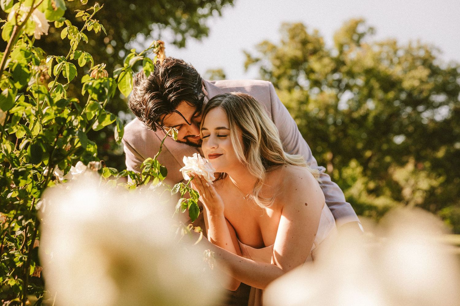 A romantic wedding couple embraces in golden sunlight beneath lush green trees during an outdoor ceremony.