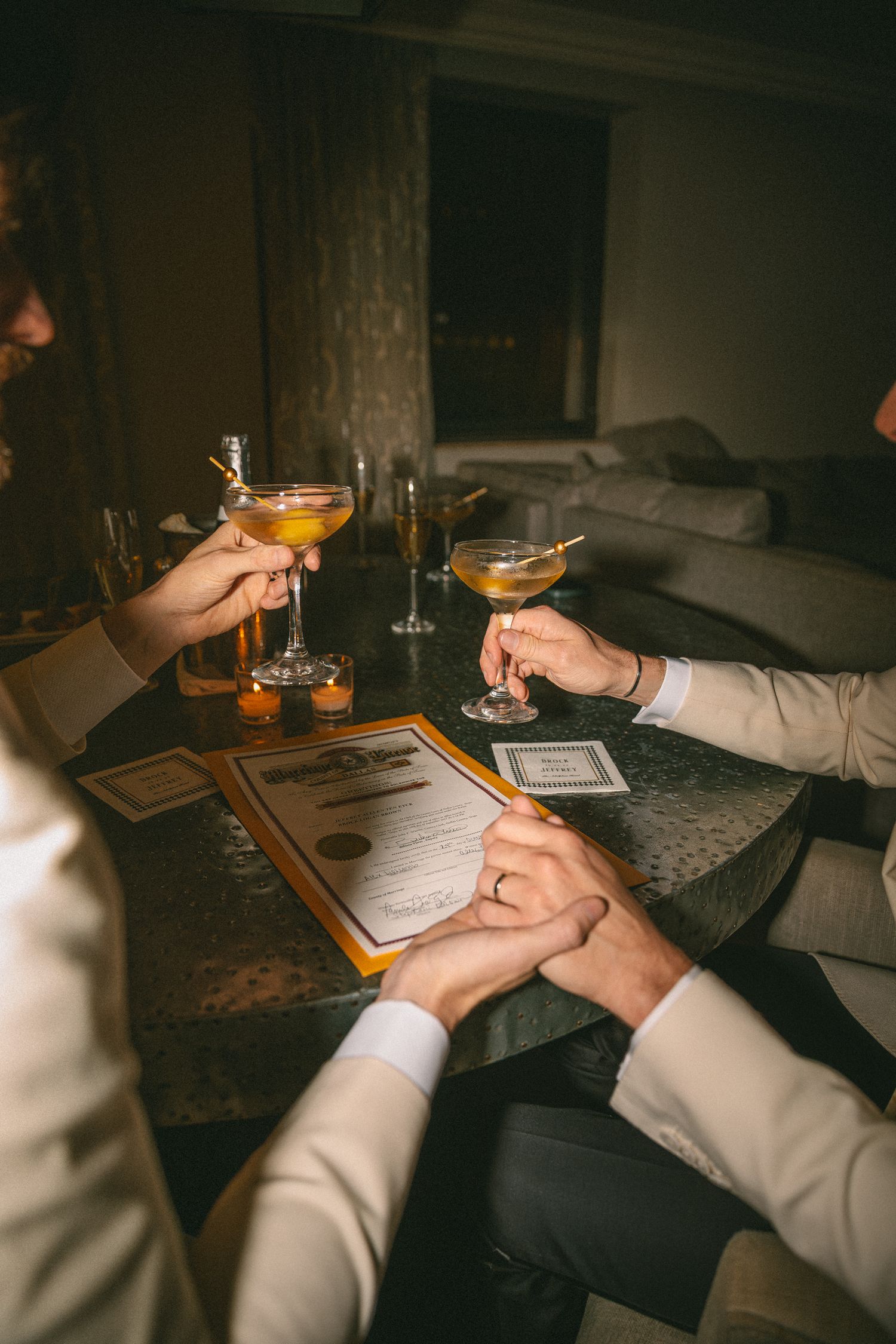 Same-sex couple holds hands and clinks cocktail glasses over marriage certificate.