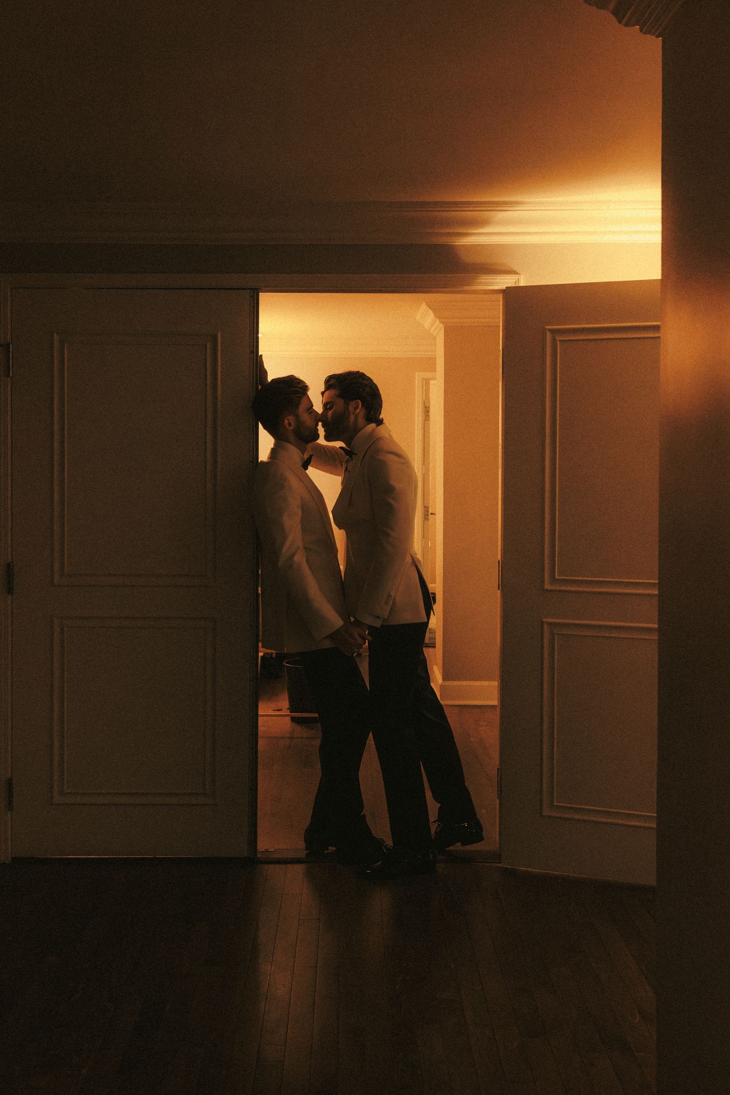 Two grooms in white tuxedos share a romantic kiss in a warmly lit doorway on their wedding day.