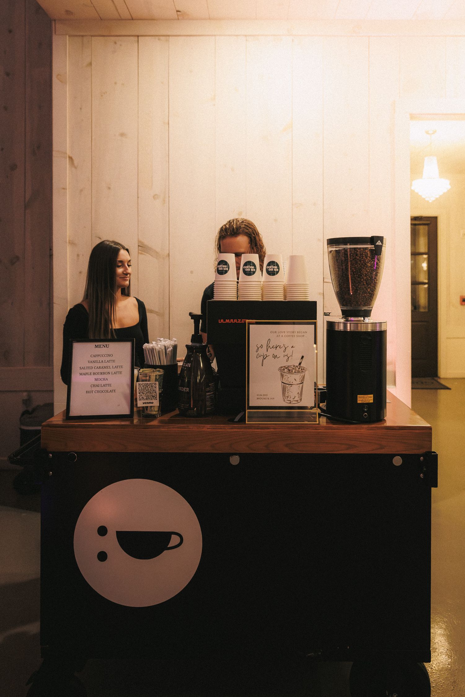 Two baristas in black standing behind a coffee bar station at a wedding reception.