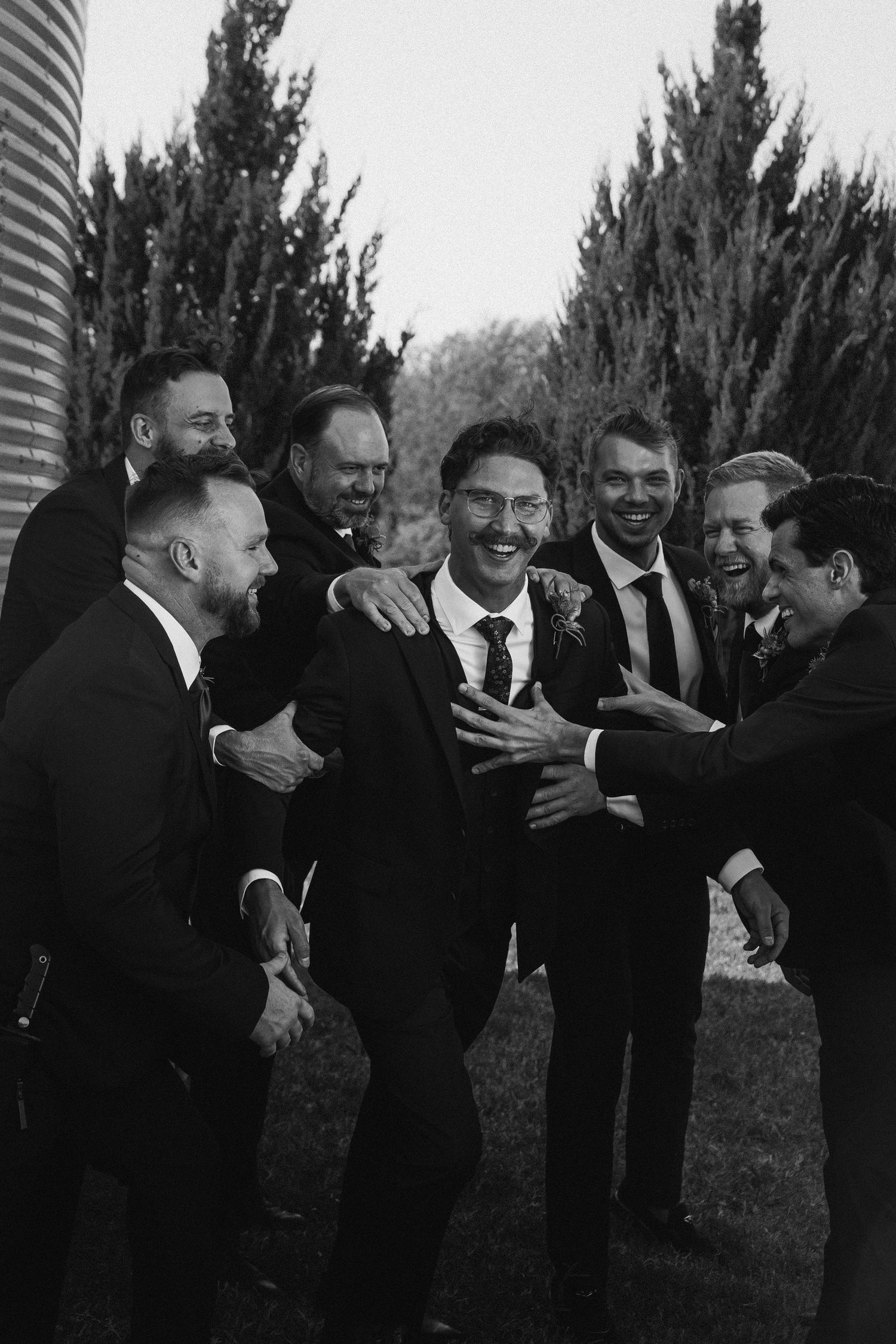 Black and white photo of a groom laughing with his groomsmen outdoors near a grain silo.