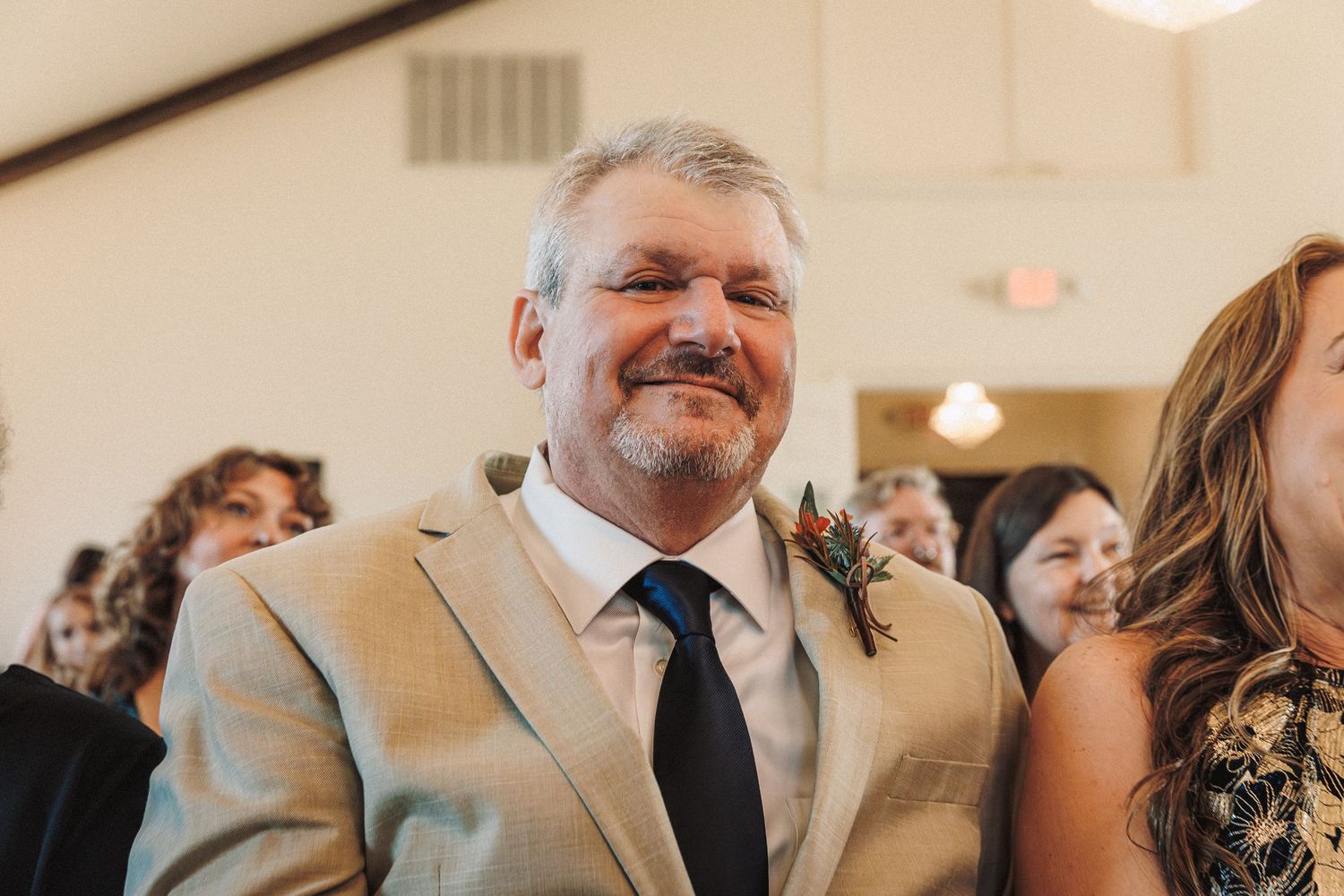 Emotional father smiling proudly in tan suit with boutonniere during wedding ceremony.