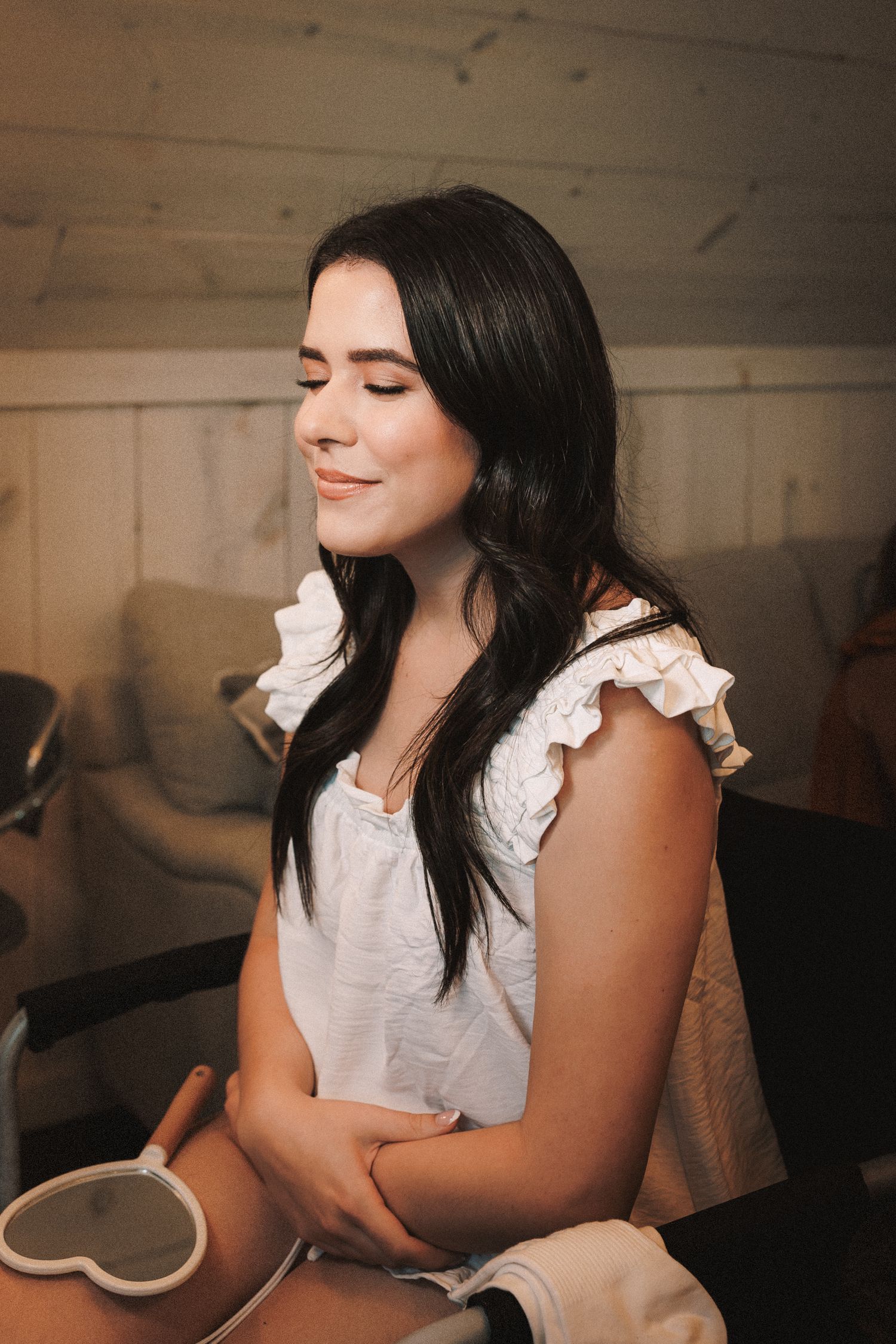 Young woman with dark hair in white ruffled top smiling with eyes closed holding heart mirror.