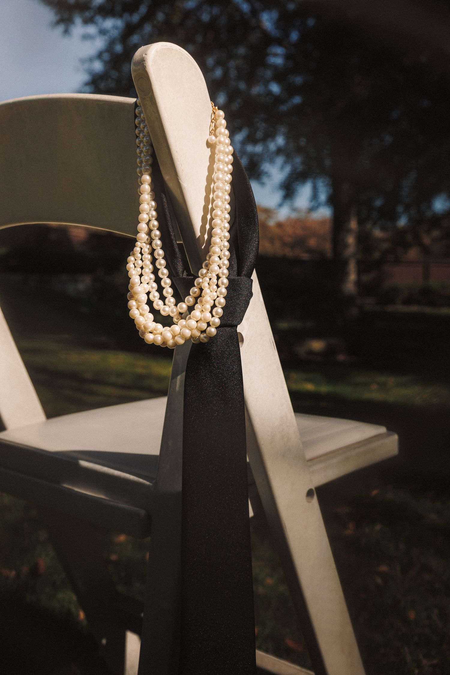 Pearl necklace draped over white folding chair with black ribbon outdoors at elegant garden wedding ceremony.