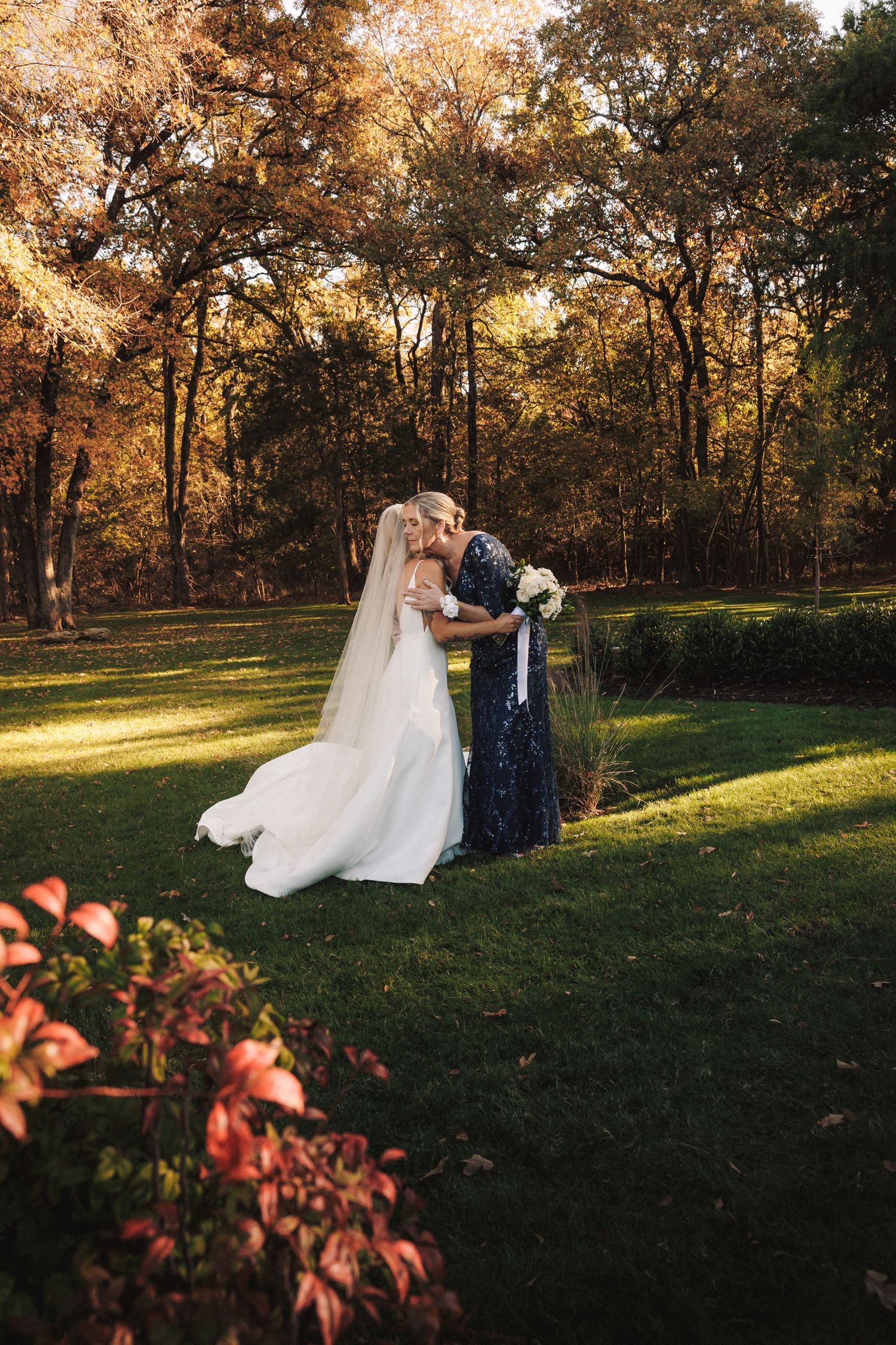 Bride in white gown and veil embraces woman in navy dress in autumn garden setting.