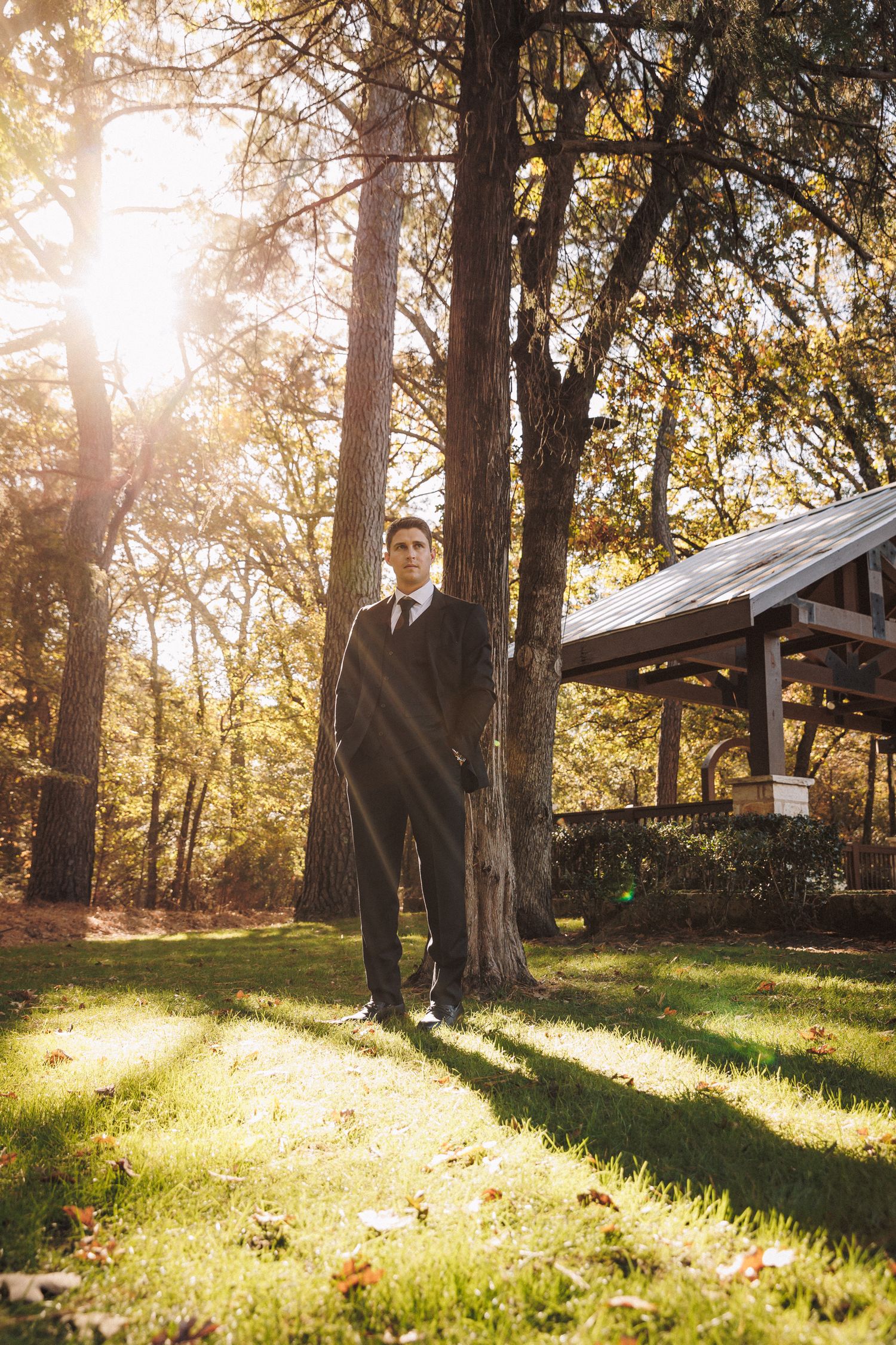 Groom in black suit standing by tree in sunlit park with pavilion background.