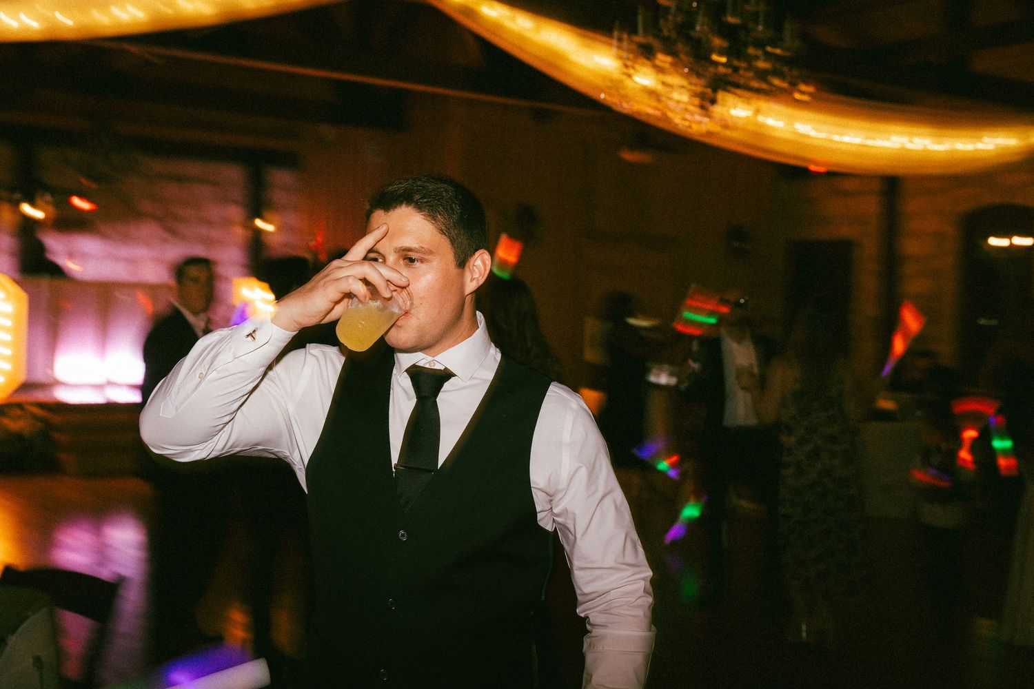 Male wedding guest drinking beer on dance floor with colorful lights at reception.
