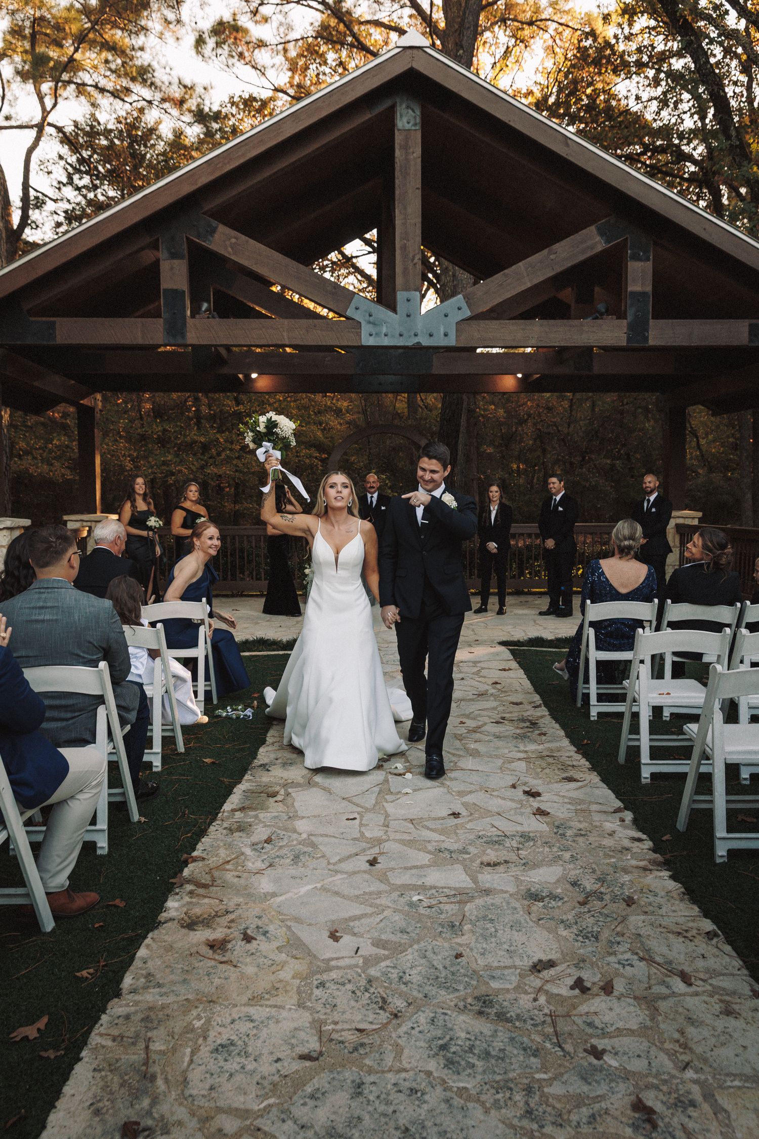 Newlyweds celebrate recessional walk down stone aisle under wooden pavilion at outdoor ceremony.