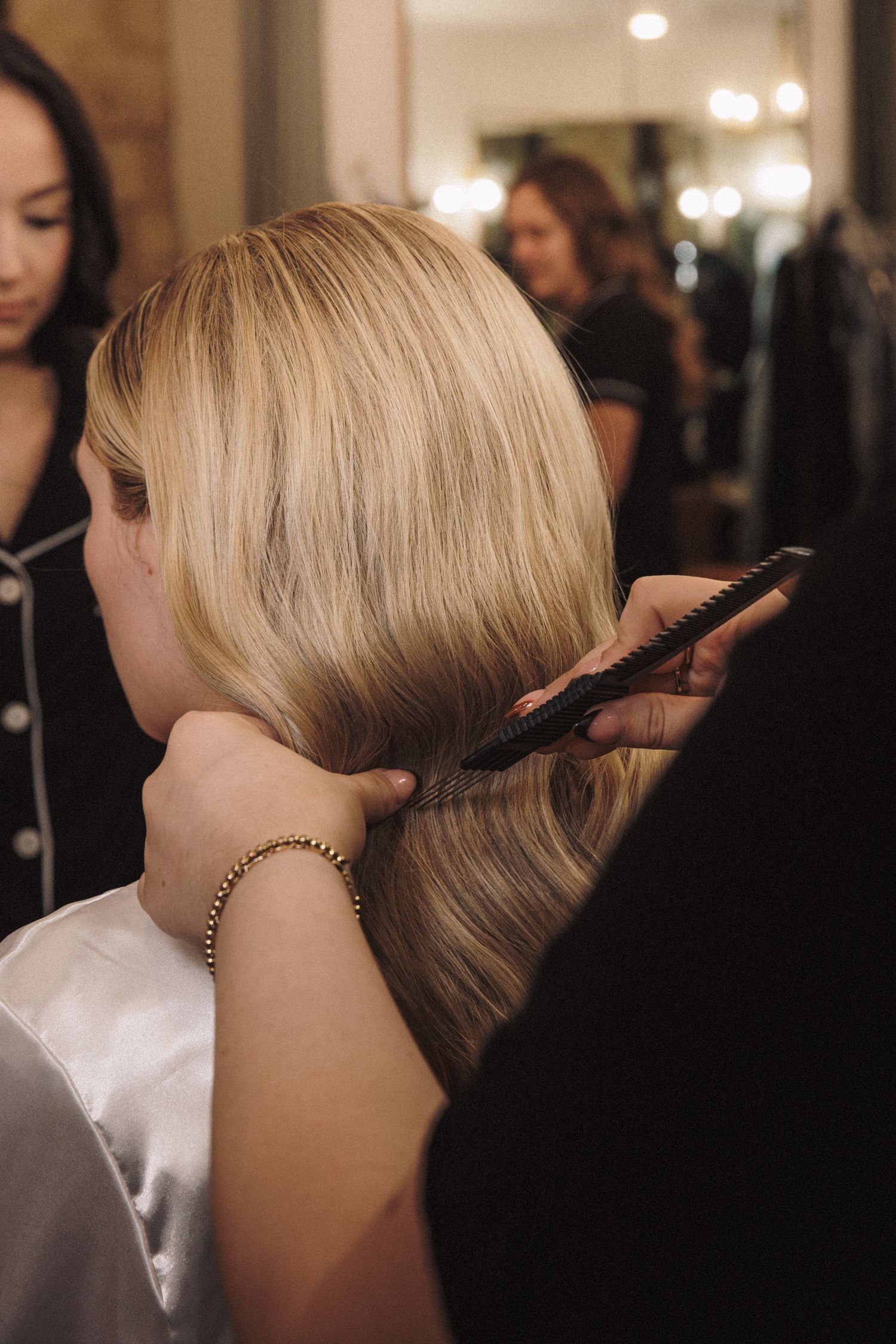 Hair stylist brushing and styling blonde bride's sleek updo hairstyle during wedding day preparations.