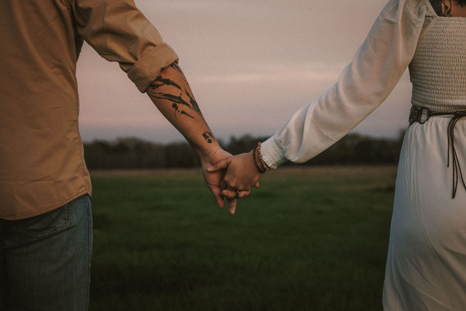 Close-up of a couple holding hands in a field at dusk, man with forearm tattoos wearing a tan jacket.
