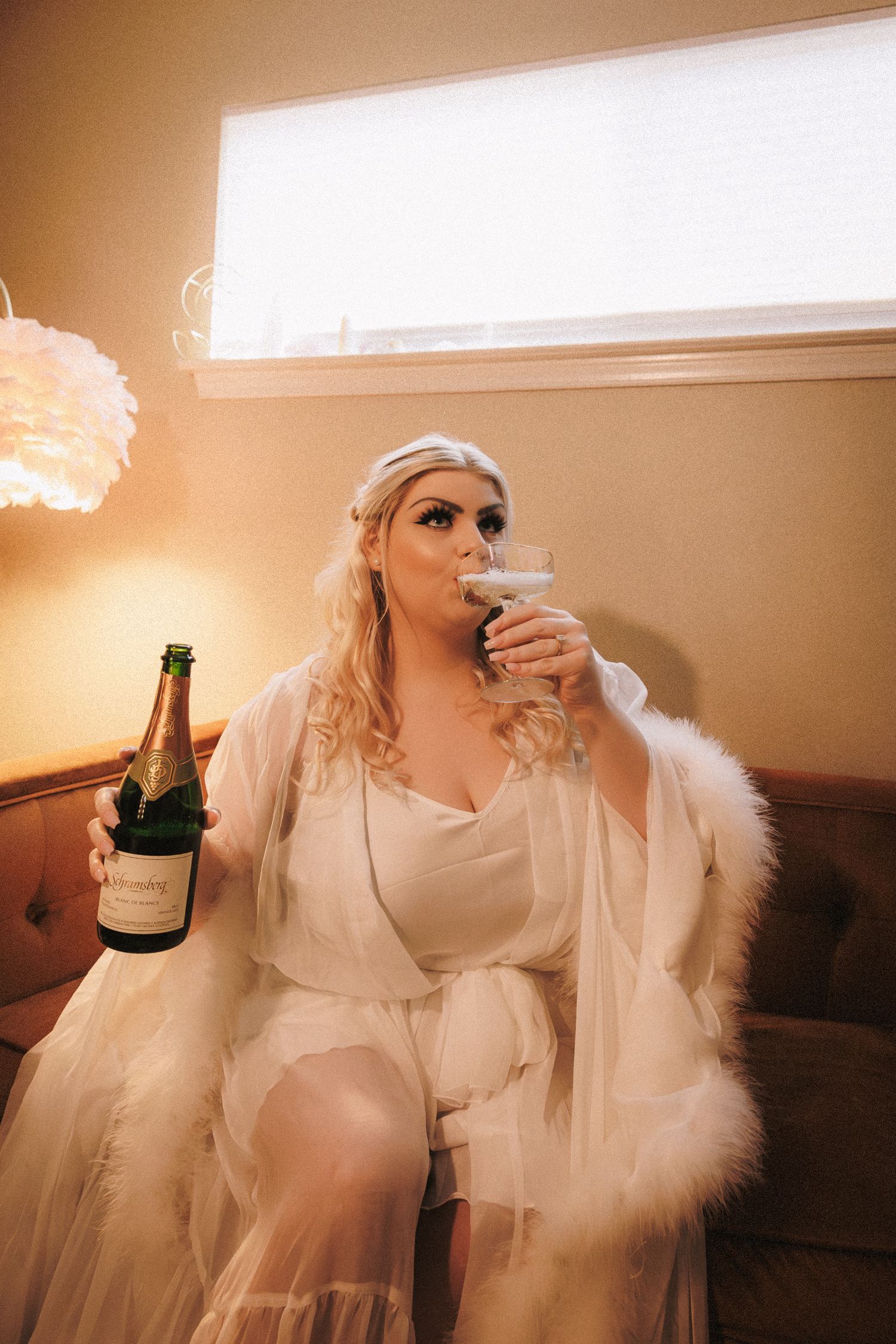 Bride in white feather robe drinking champagne from a coupe glass holding a bottle.