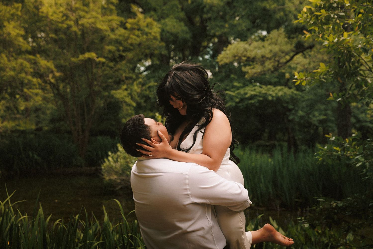 A couple shares an intimate embrace while standing outdoors in a lush garden setting during golden hour.