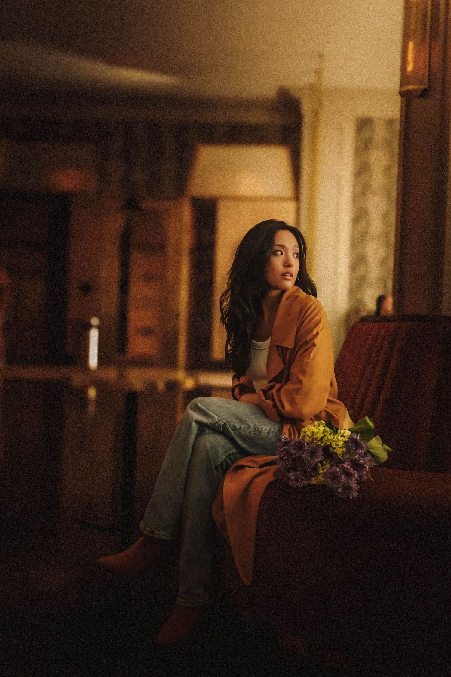 Moody portrait shot of a person in orange sweater sitting with flowers on a couch in a dimly lit rustic room.