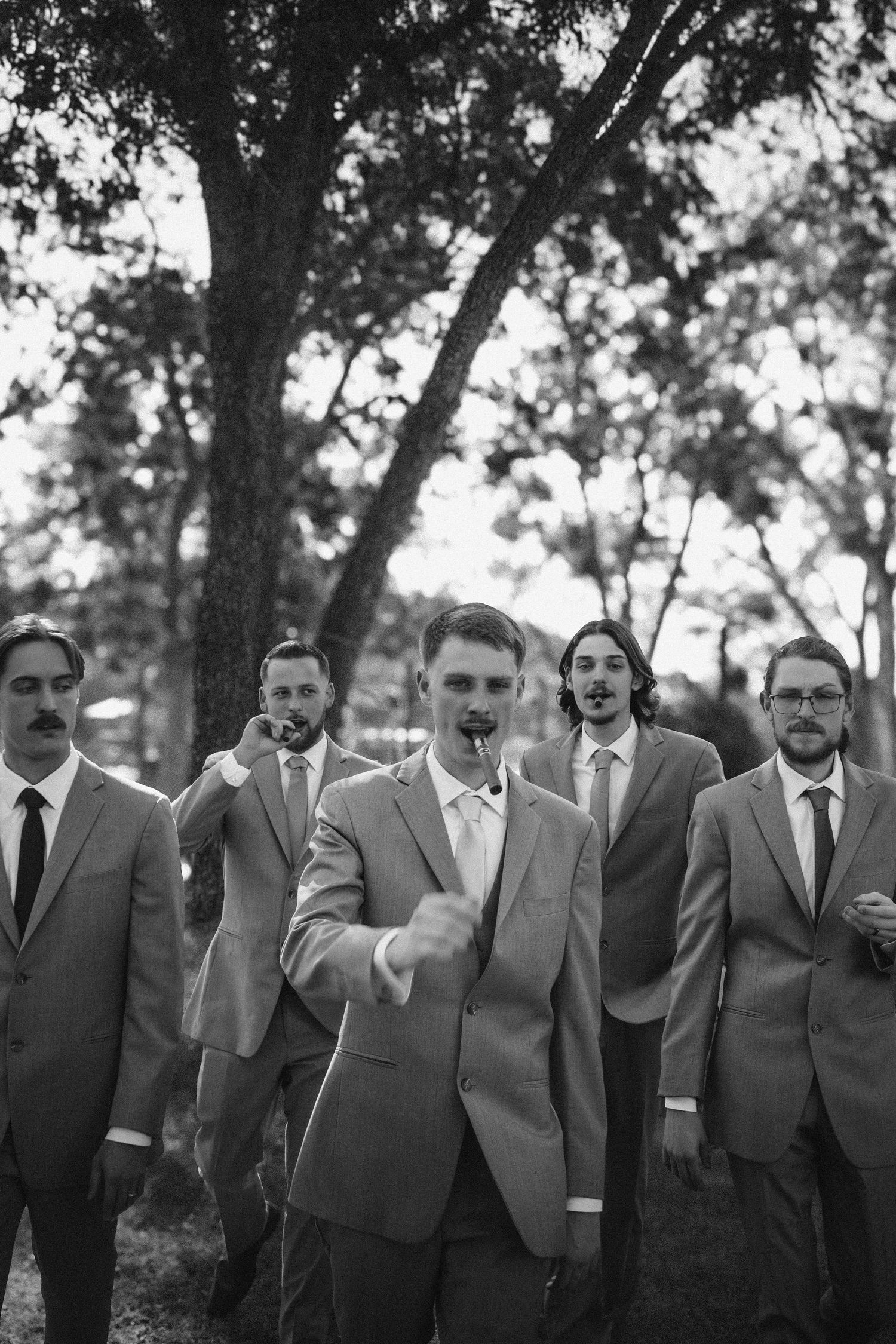 A group of groomsmen in matching gray suits pose together outdoors in a black and white photo.