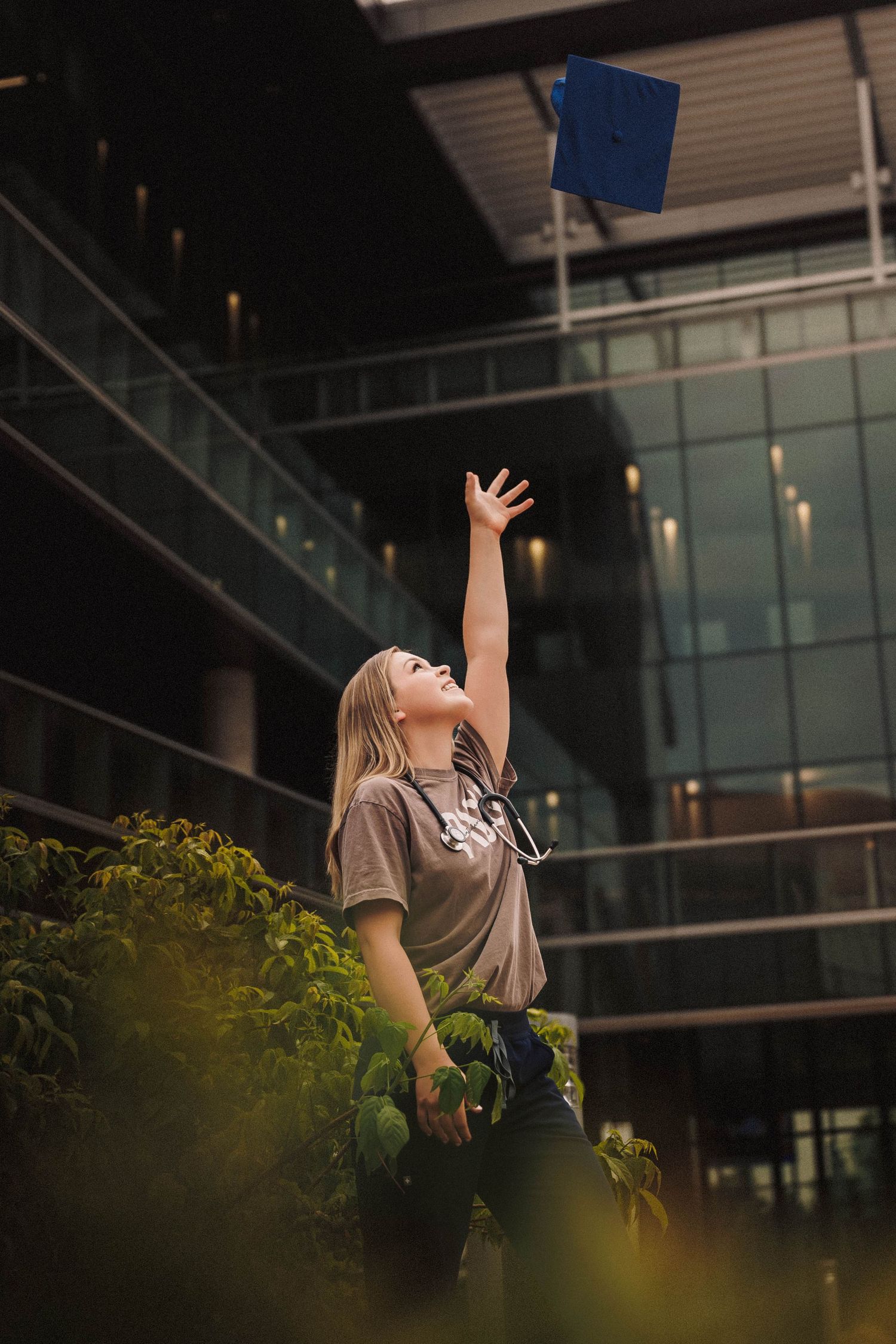 A person in a beige shirt poses with outstretched arms against a modern building exterior with greenery in the foreground.
