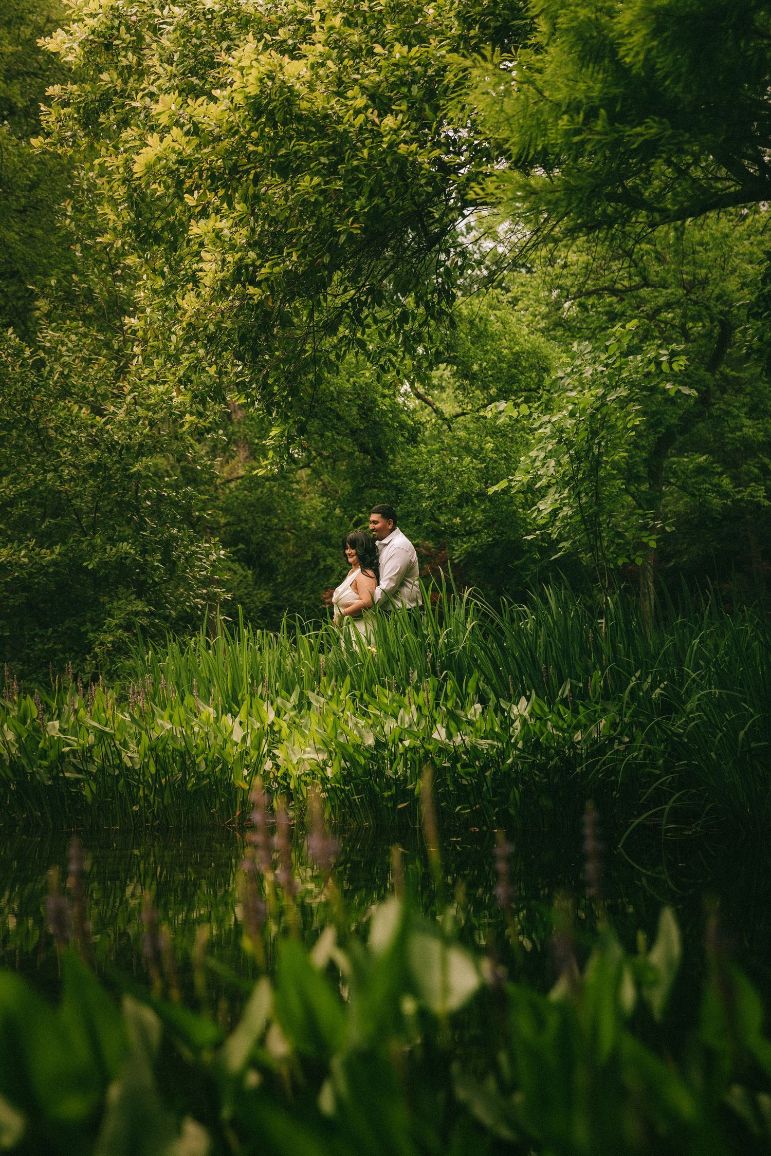 A romantic couple enjoys an intimate moment by a tranquil pond surrounded by lush green foliage on a summer day.