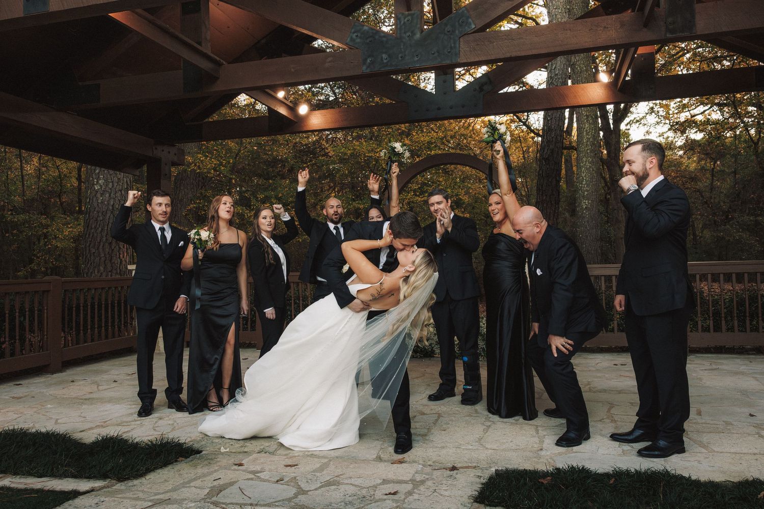 Wedding party celebrates as groom dips bride under rustic outdoor pavilion at night.